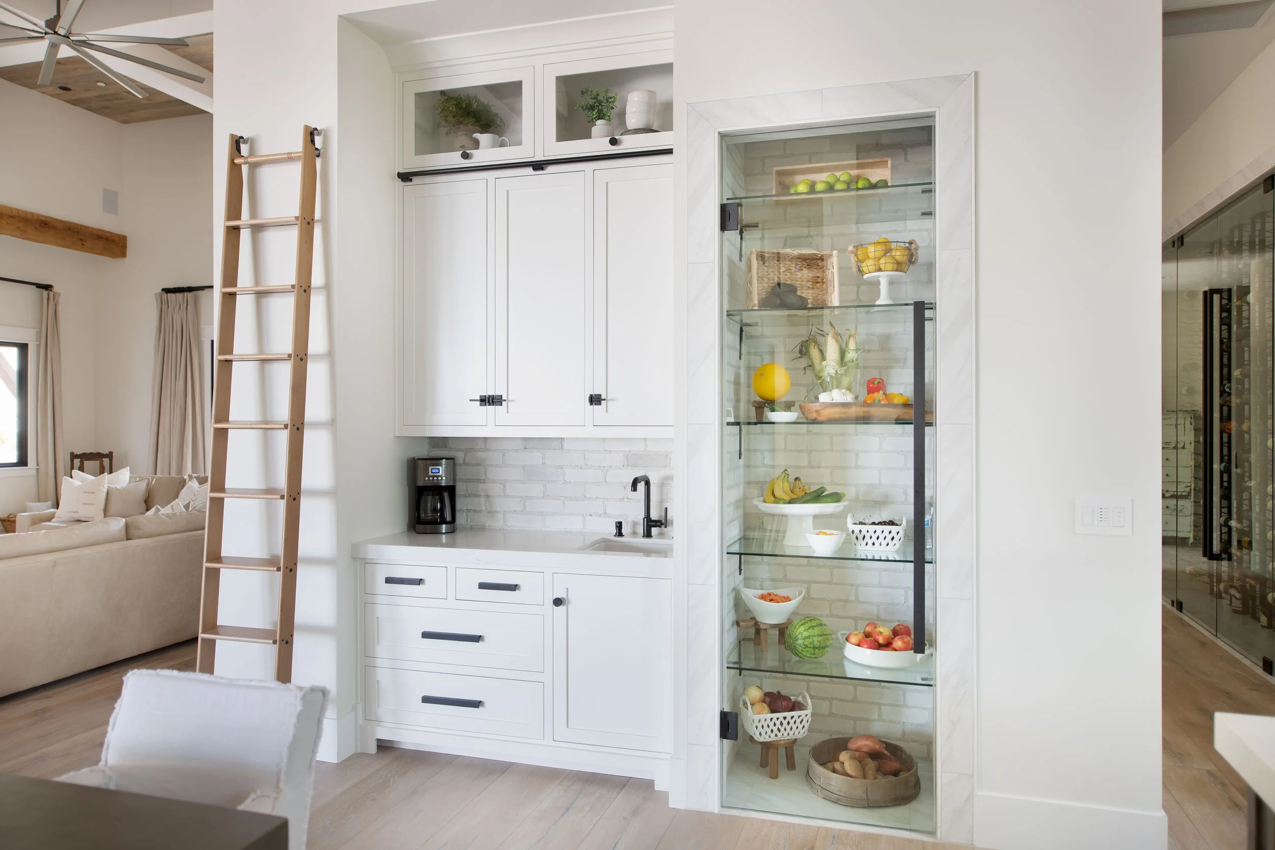 Modern kitchen area with white cabinetry, a glass display case filled with green apples, lemons, bananas, root vegetables, and other fresh produce. Part of a living room with a beige sofa and wooden beams visible in the background.