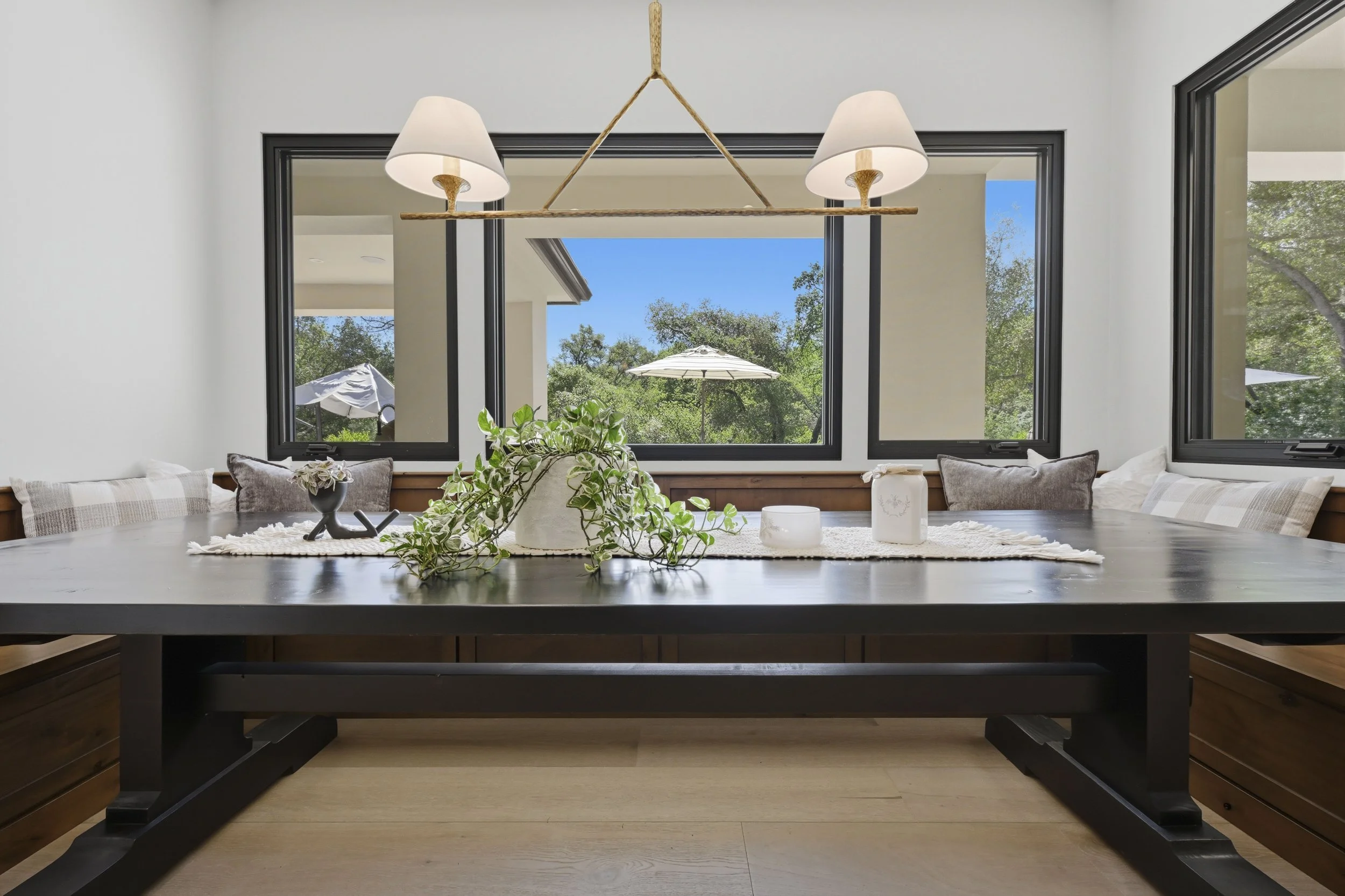 Dining room with a large black table, a potted plant and decorative items, three large windows showing a sunny outdoor patio with a white umbrella and trees, and a modern hanging light fixture with two lampshades.