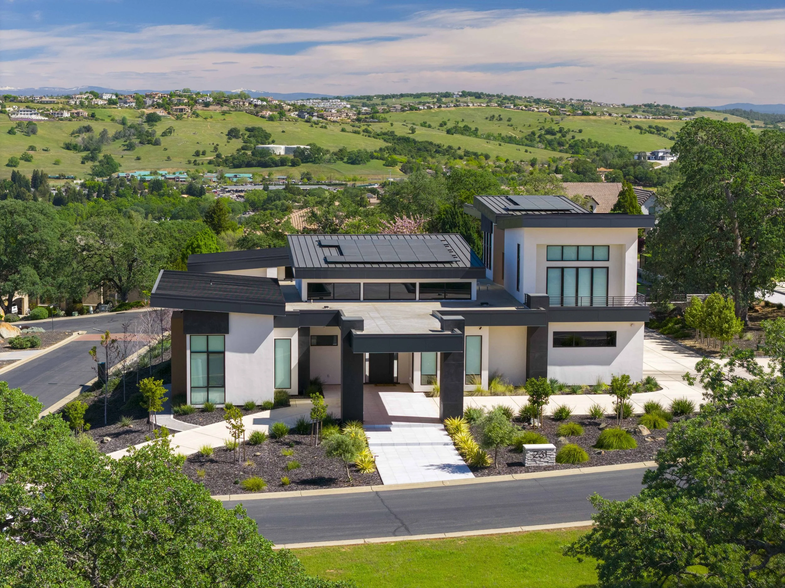 Modern white house with flat roof and solar panels, surrounded by landscaped garden with small trees and bushes, adjacent to a street.