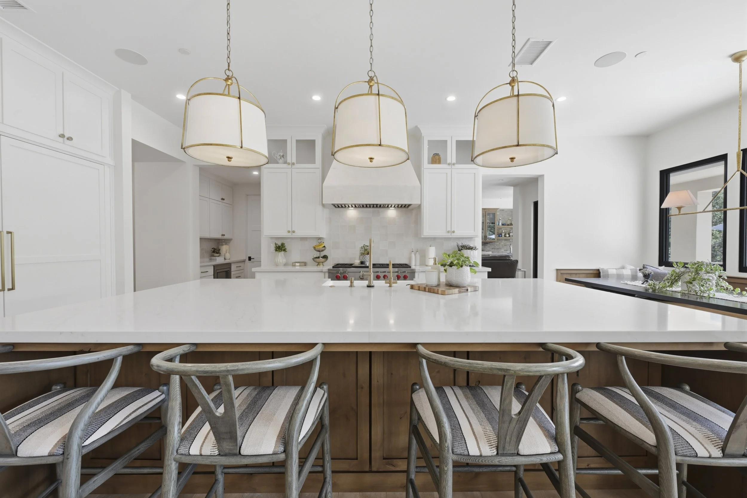 A modern kitchen with white cabinets, a white marble island, and three large pendant lights hanging above. Brown and beige striped chairs are arranged at the island. The background shows a window and a living space with a mirror and greenery.