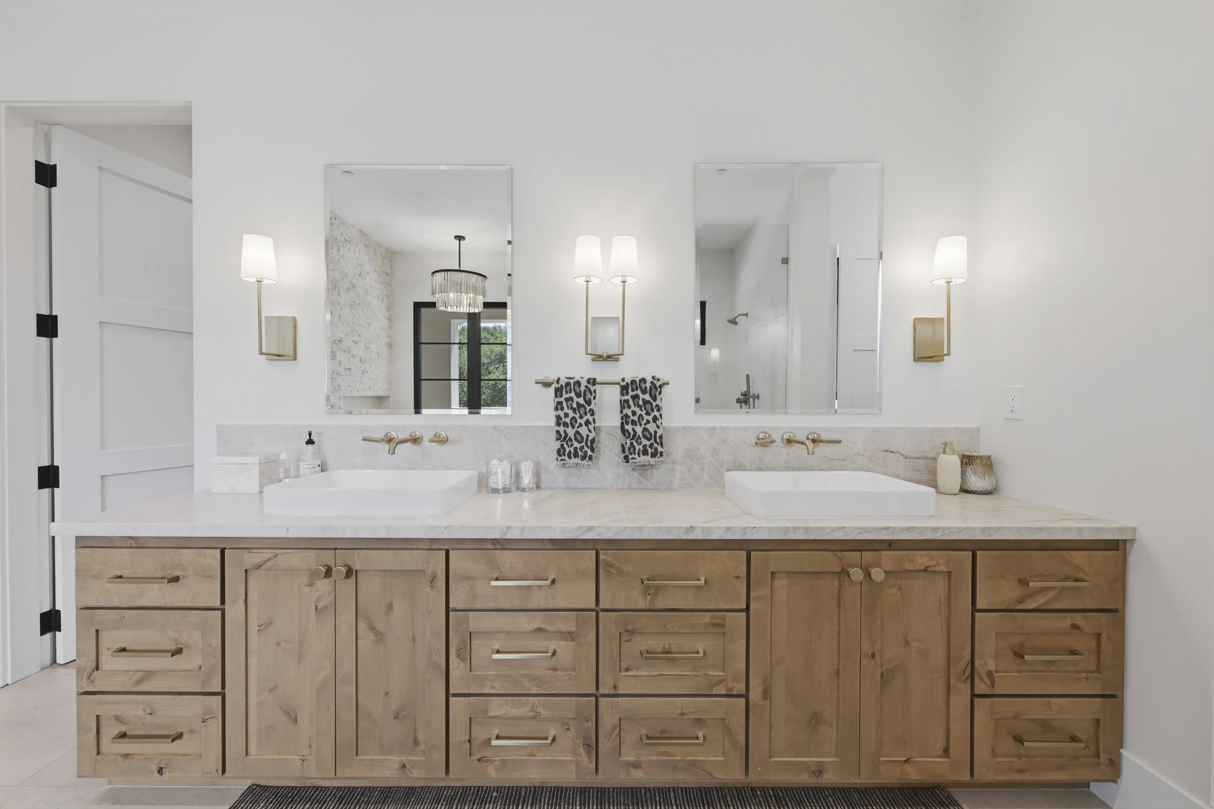 Bathroom vanity with a marble countertop, dual square sinks, wall-mounted faucets, and wooden cabinets, with mirrors, wall sconces, and decorative items.