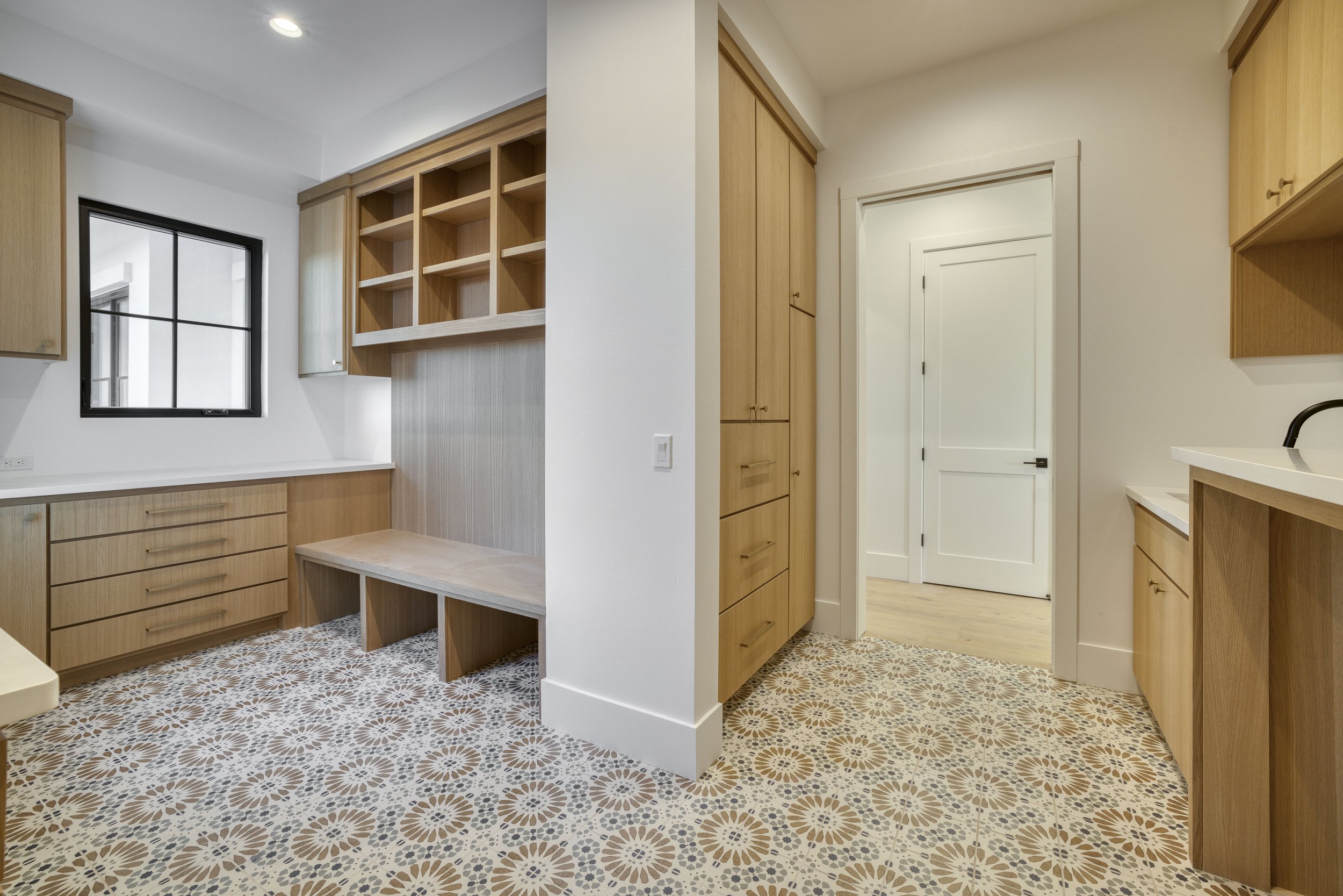 Interior view of a room with wooden cabinetry, a window with black framing, patterned tile flooring, and a doorway leading to another room.