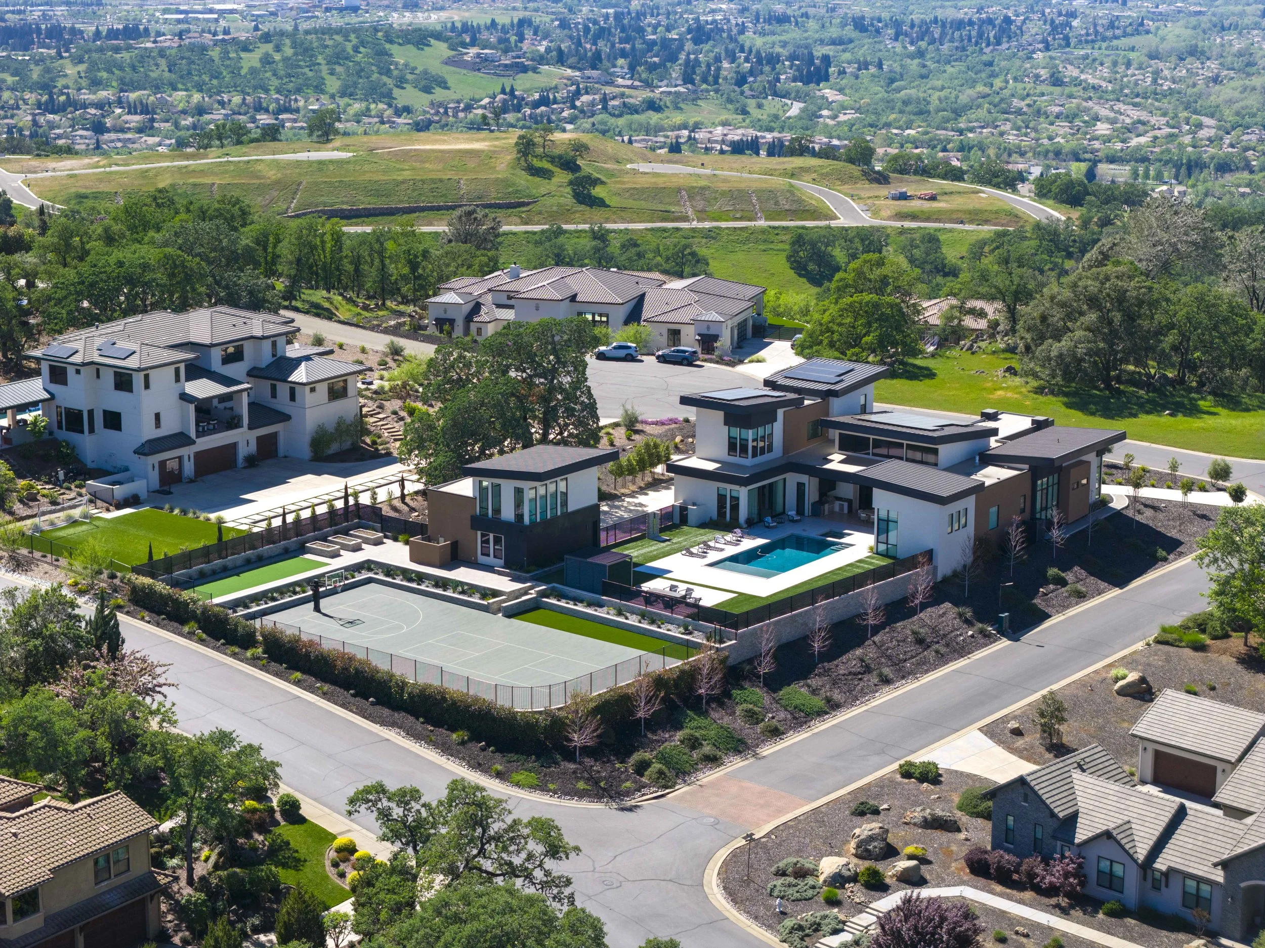 An aerial view of a residential neighborhood with modern houses, green lawns, and a sports court, surrounded by trees and hilly terrain.