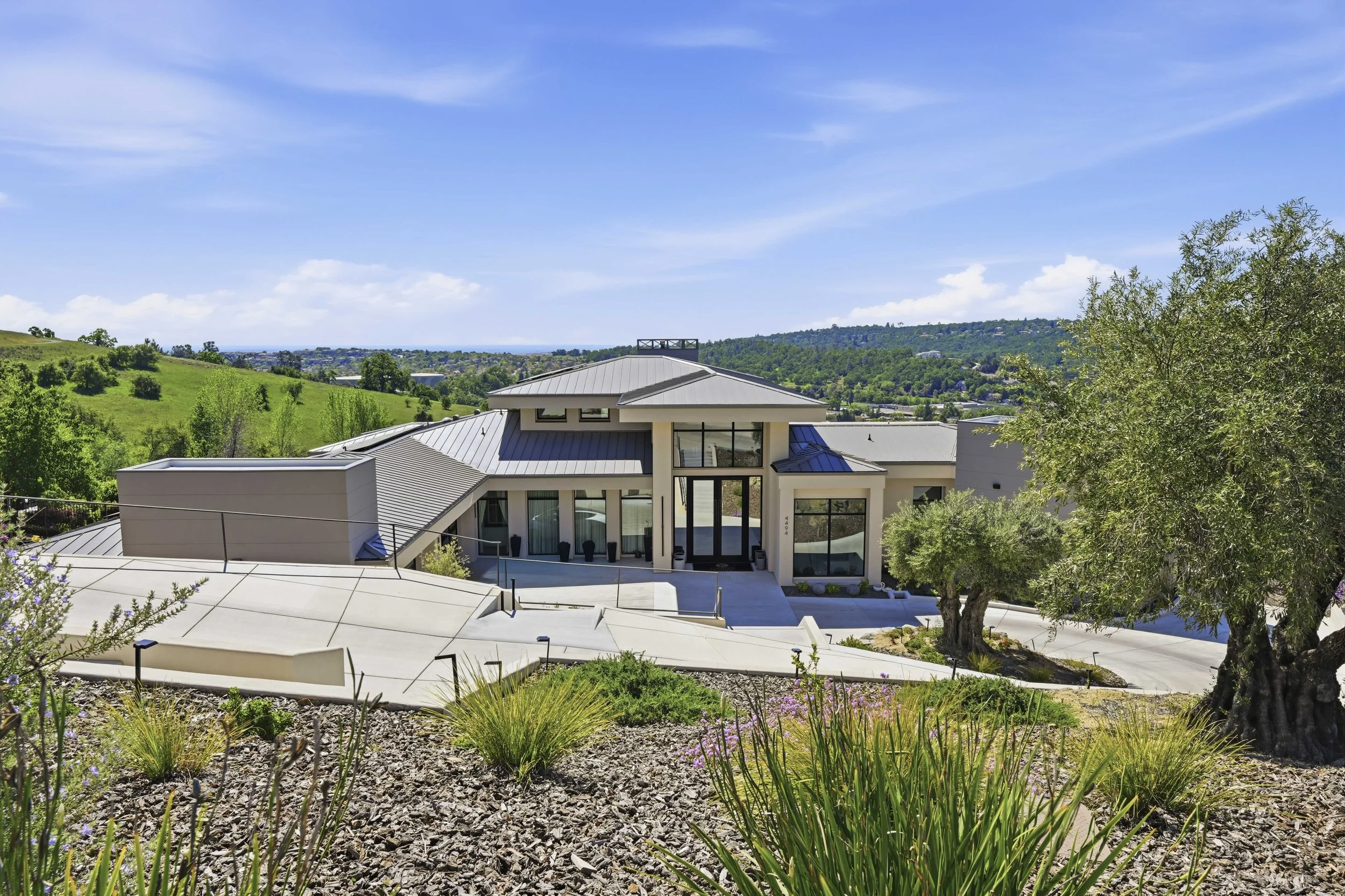Modern house with large windows and metal roof, surrounded by a landscaped yard with trees, plants, and hills in the background under a blue sky.