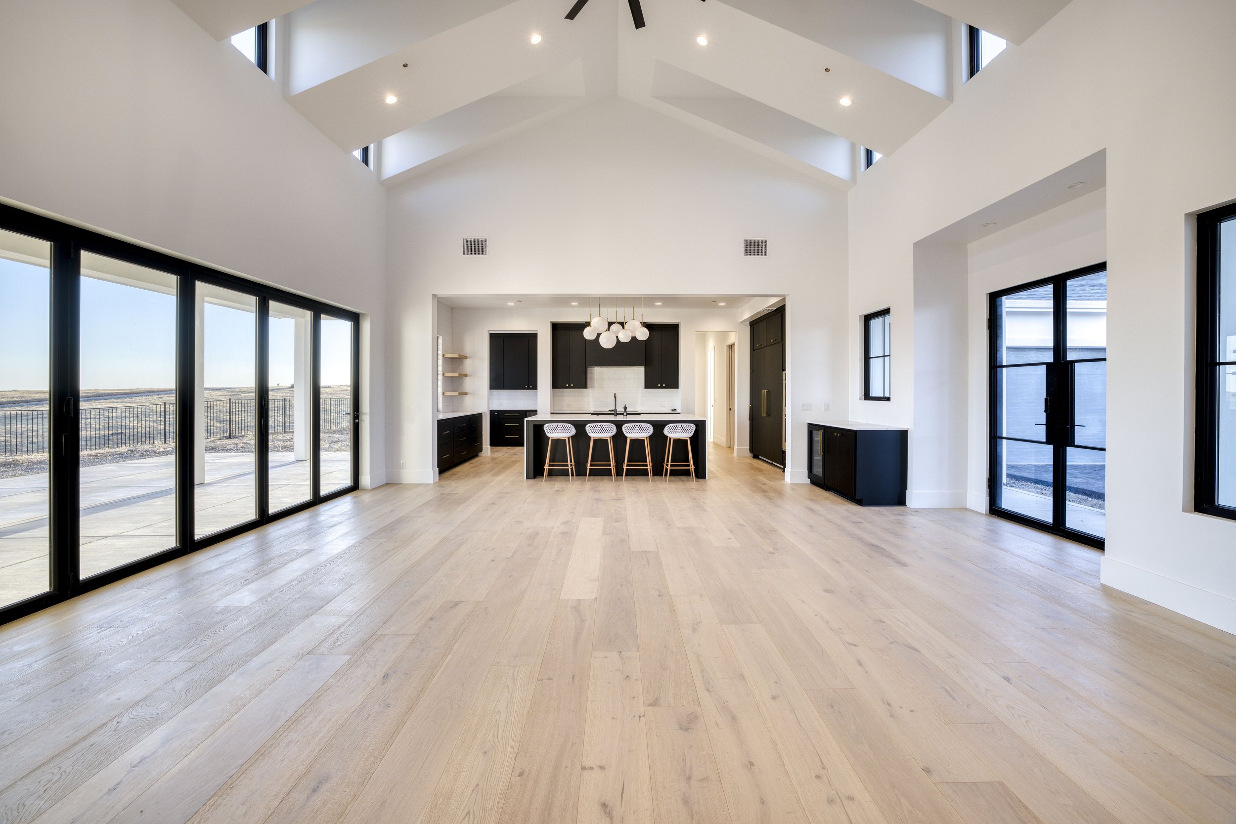 Spacious modern living room with large black corner-to-corner windows, white walls, light wood floors, and a kitchen area with black cabinets, white backsplash, and a breakfast bar with three white chairs. Ceiling lights and a ceiling fan are visible
