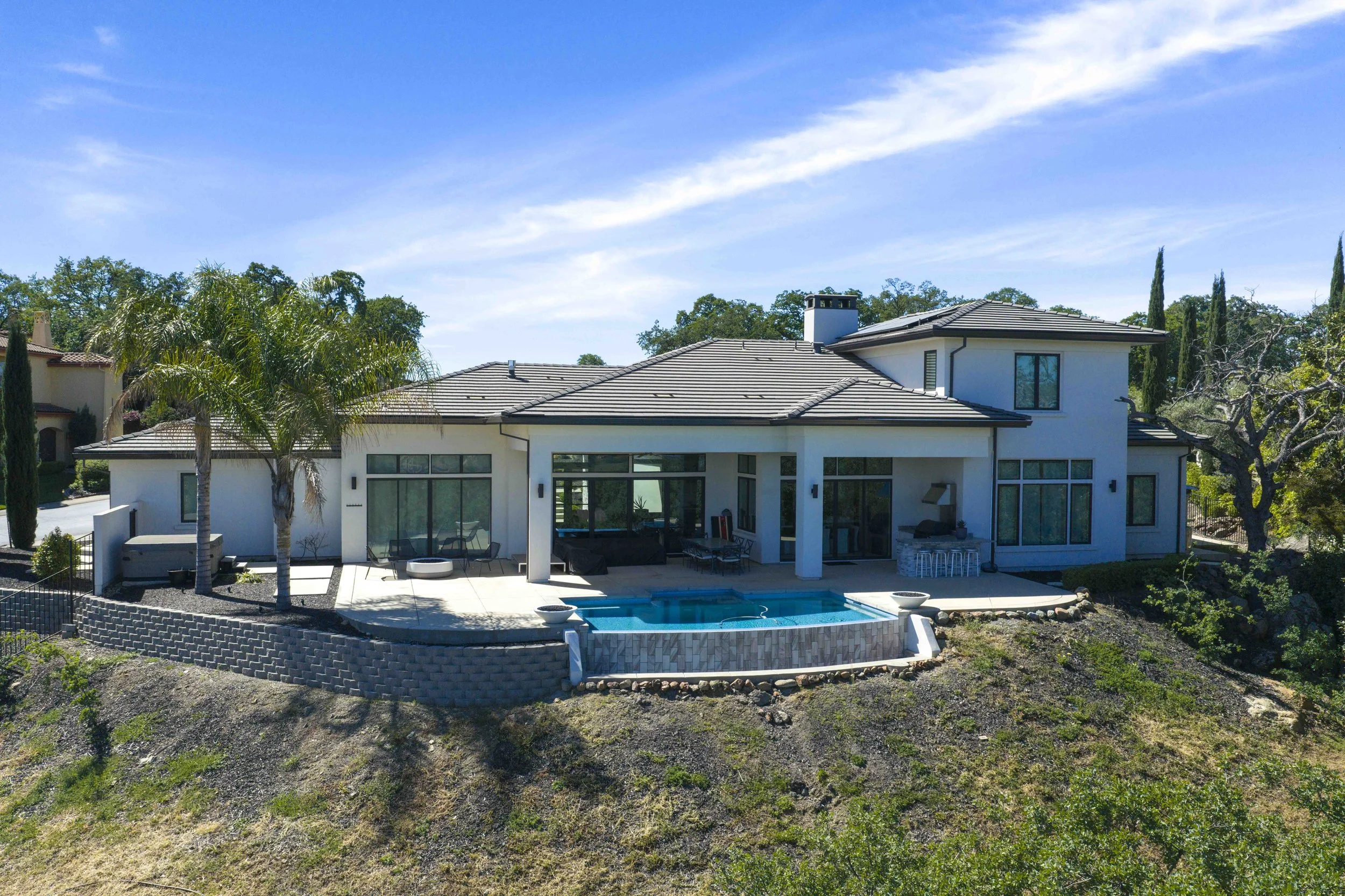 Modern white house with large windows, patio, swimming pool, and a backyard garden with trees, shrubs, and a tiered stone retaining wall under a partly cloudy blue sky.
