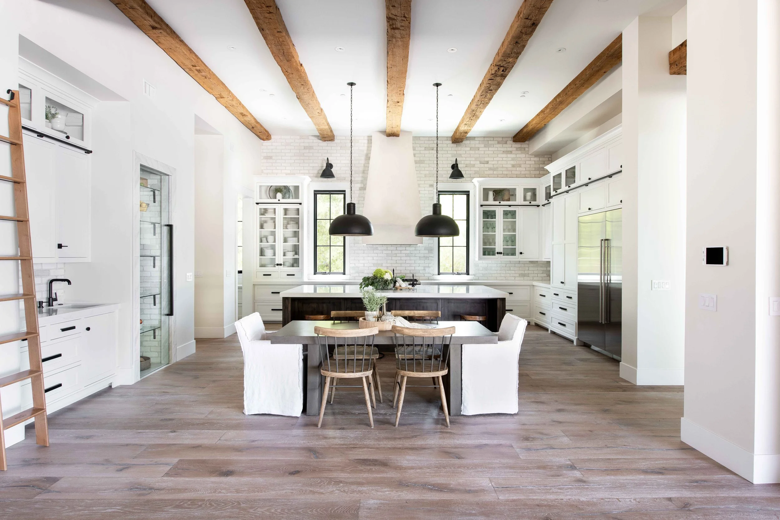 Modern kitchen with white cabinets, black hardware, and a central island. Wooden ceiling beams, black pendant lights, and large windows with black frames. Wooden dining table with white and wooden chairs, and a wooden ladder on the left.