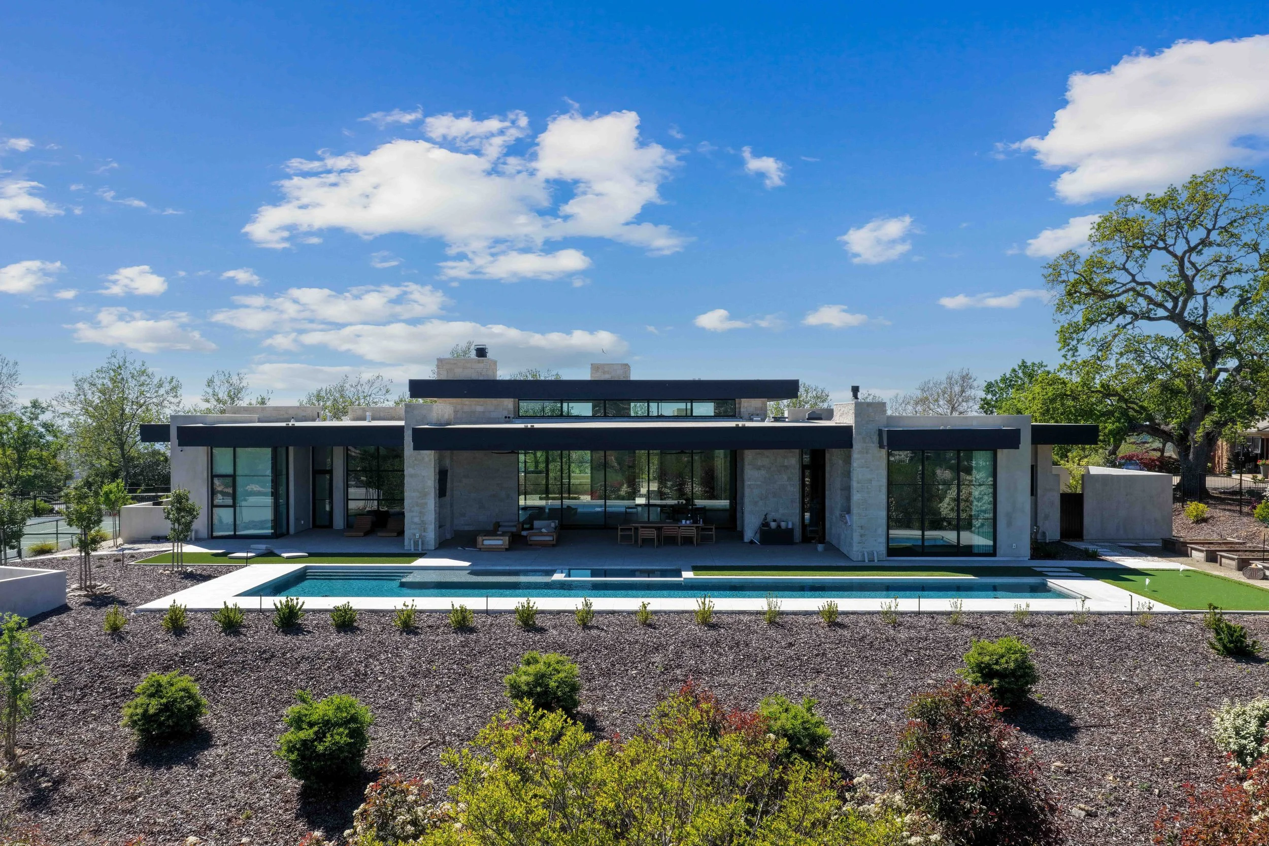 Modern house with a flat roof, large glass windows, a swimming pool, and a landscaped yard with small bushes and trees under a bright blue sky with scattered clouds.