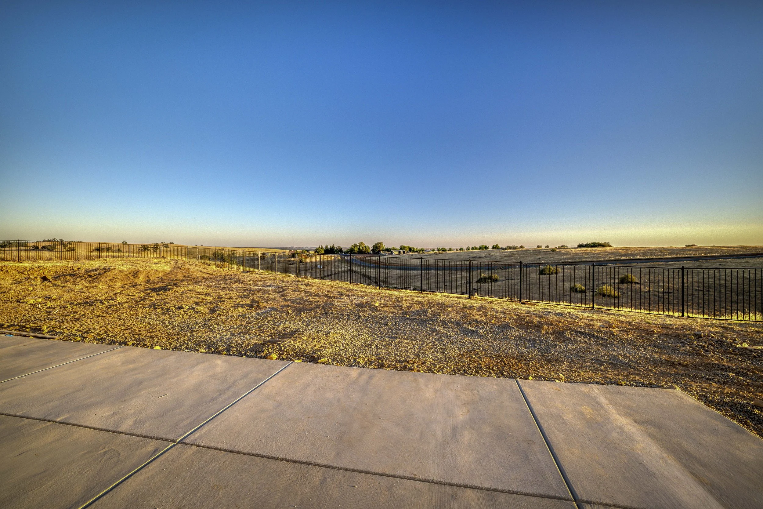 A wide open landscape with a dirt and gravel foreground, a black metal fence, and dry grassy hills under a clear blue sky.