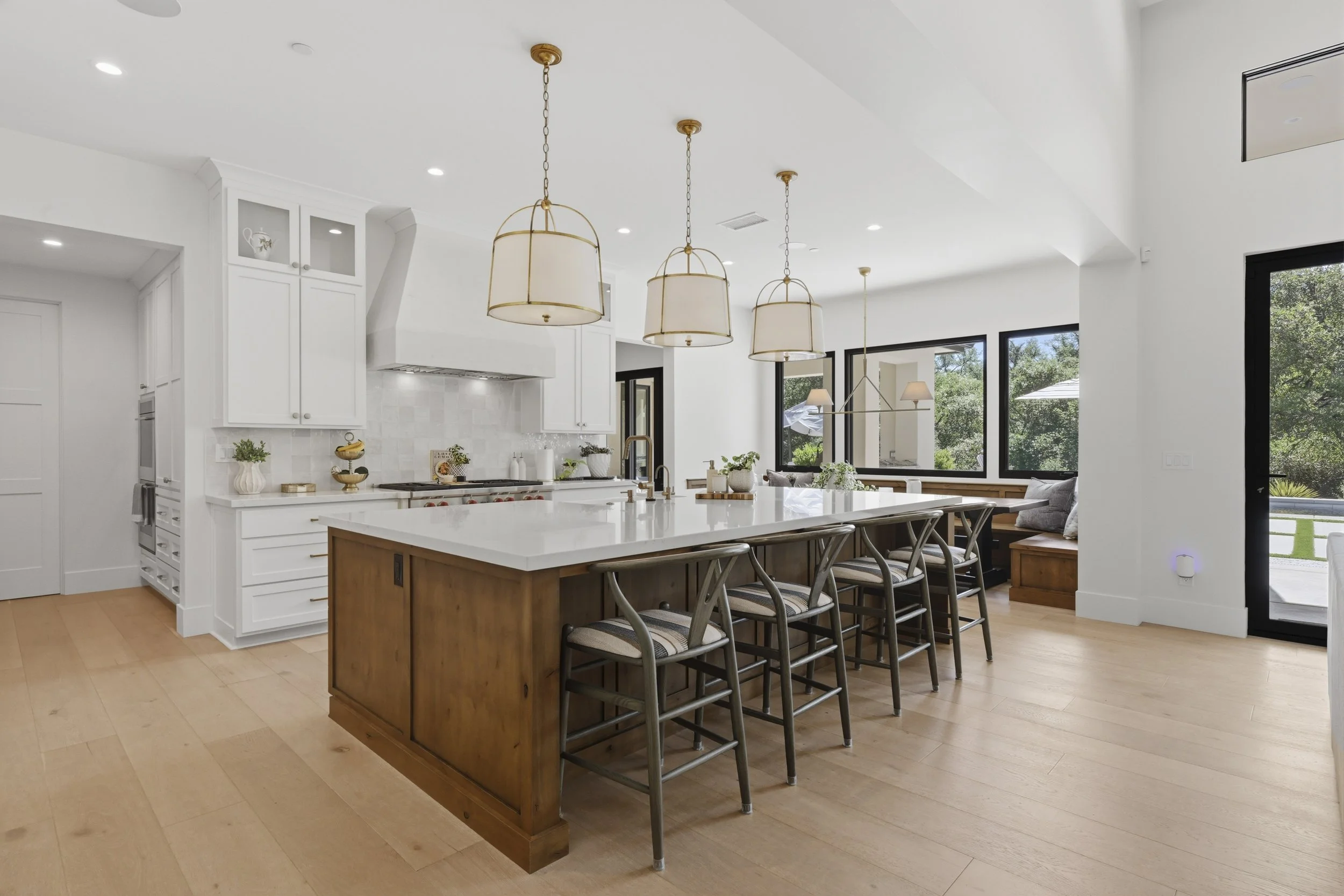 Modern kitchen with white cabinets, a large kitchen island, wooden bar stools, and large windows with outdoor greenery.