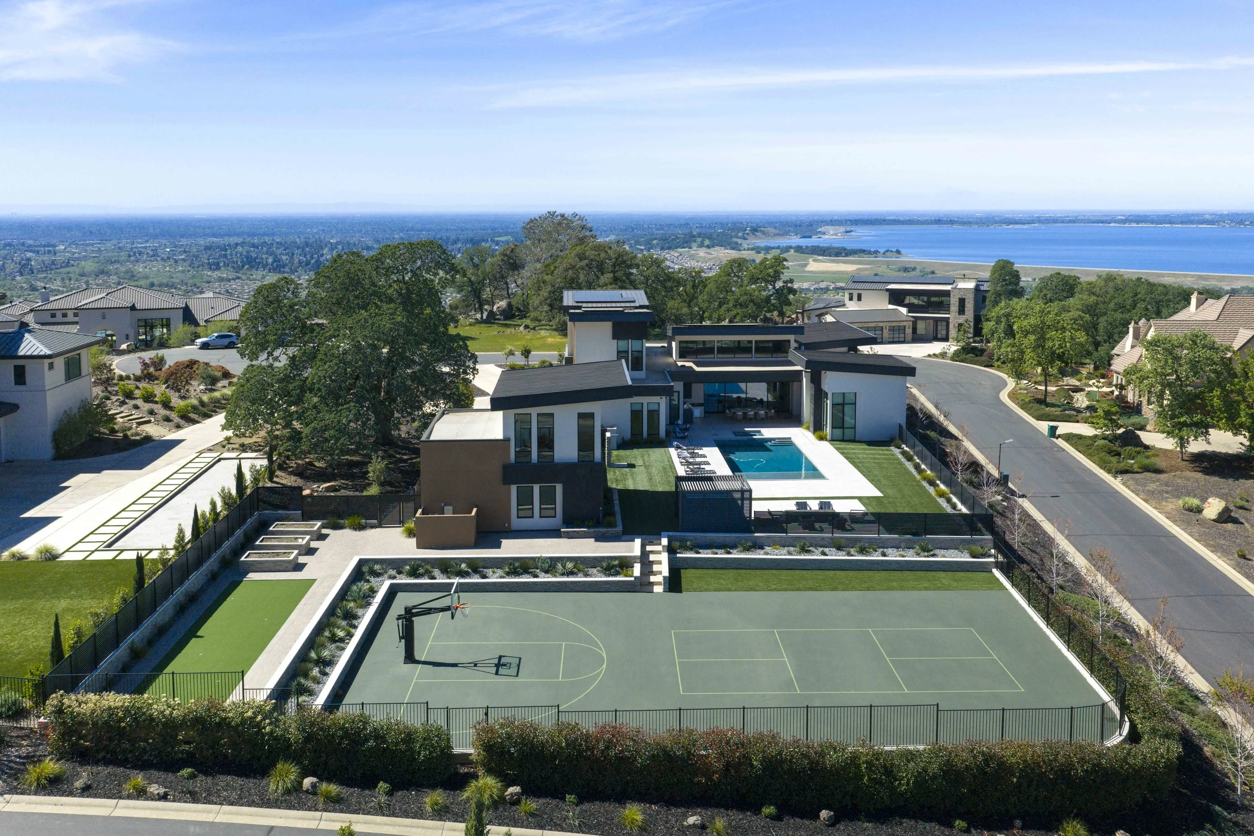 Aerial view of a modern residential neighborhood with a backyard featuring a swimming pool and a basketball court, overlooking a large body of water and distant landscape.