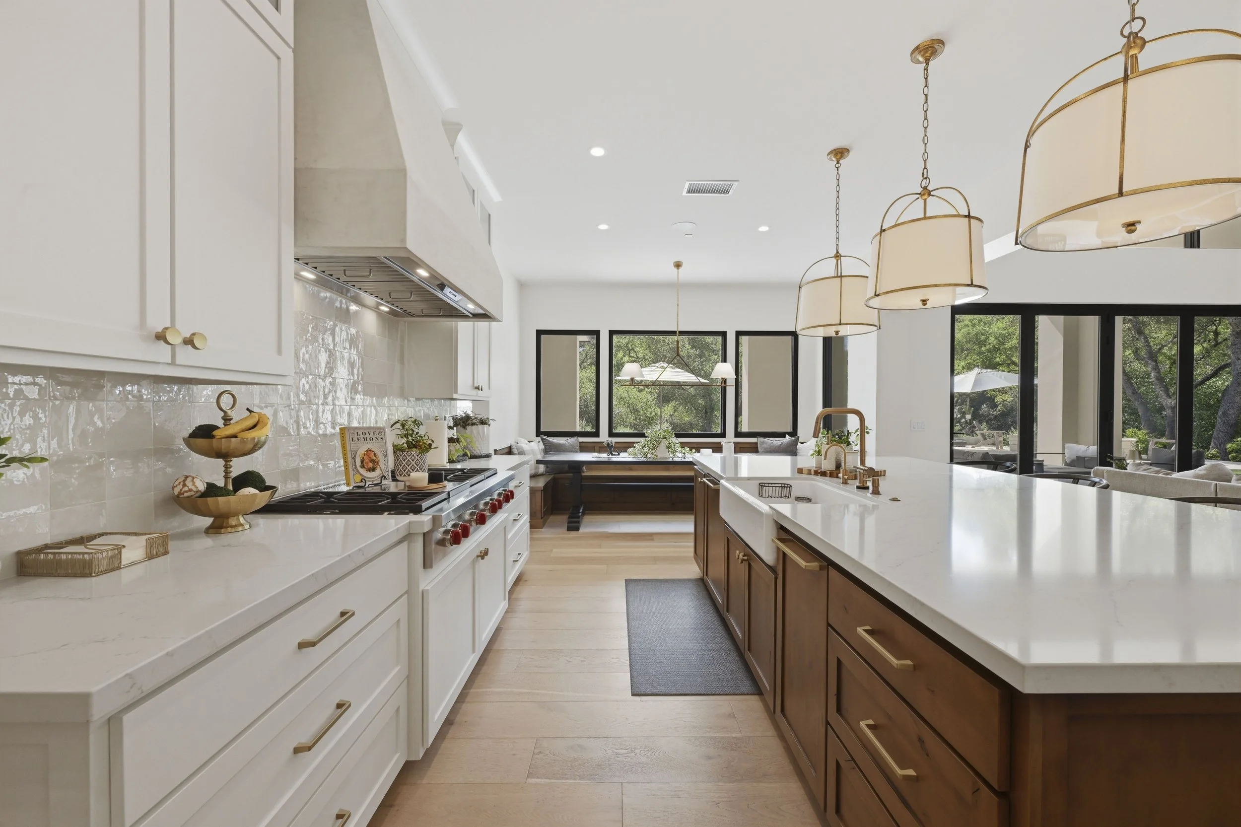 A modern kitchen with white and wooden cabinets, a white marble island, pendant lights, and large windows showing outdoor trees.