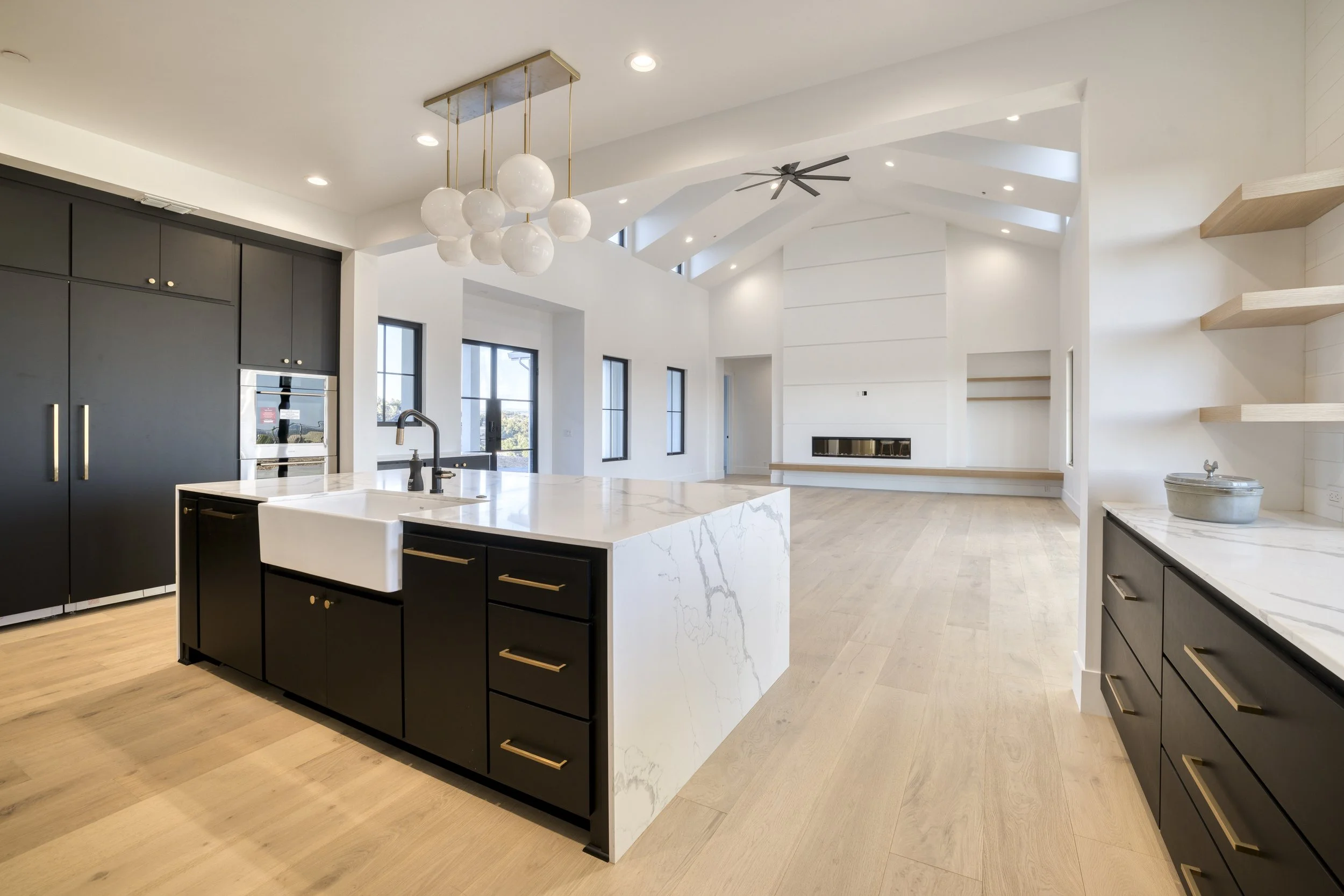 Modern open-concept kitchen with black cabinets, a large white marble island, light wood flooring, high ceilings with skylights and a ceiling fan, and large windows letting in natural light.