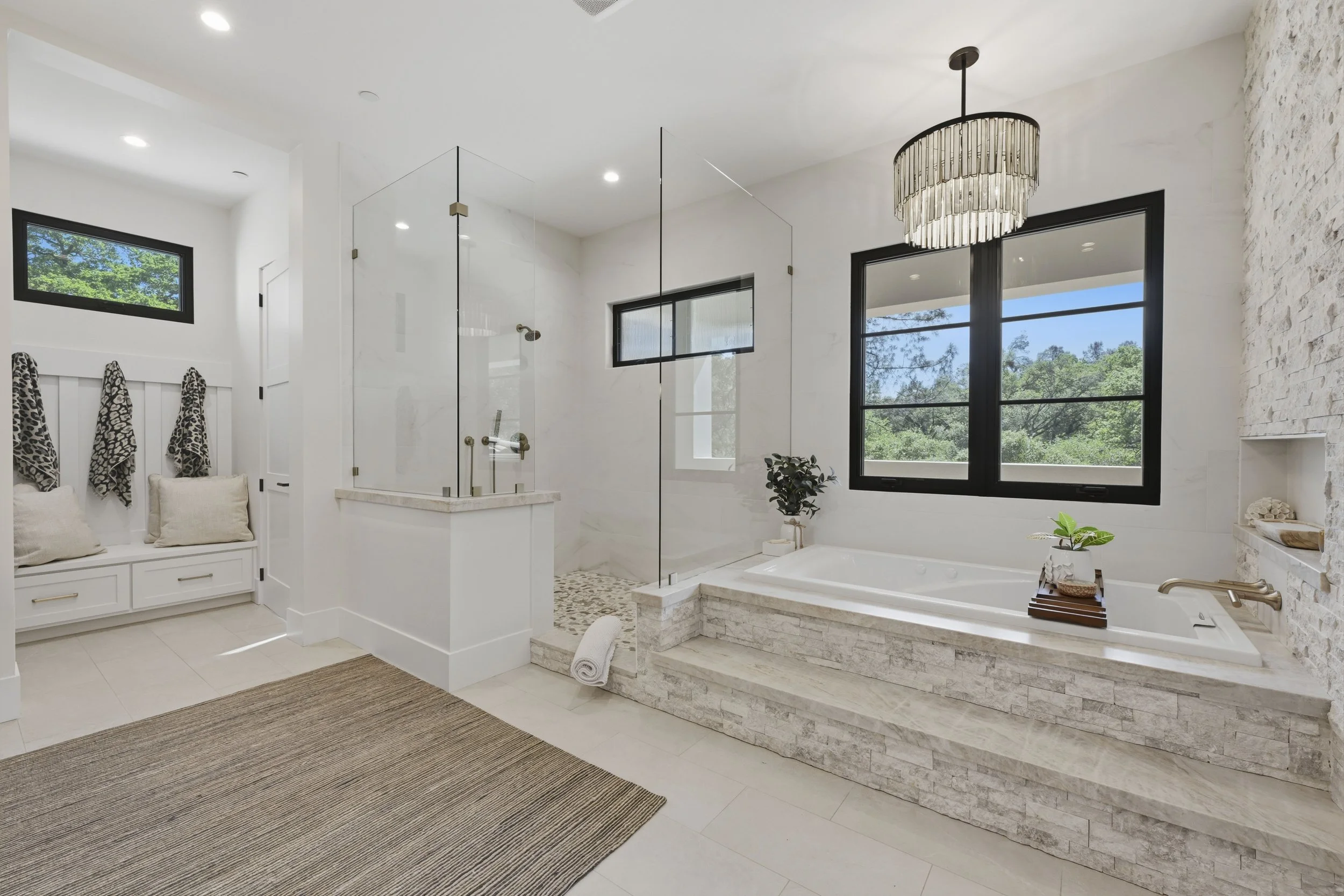 Modern bathroom with large windows, a glass-enclosed shower, bathtub with stone surround, and minimalist decor including plants and towels.