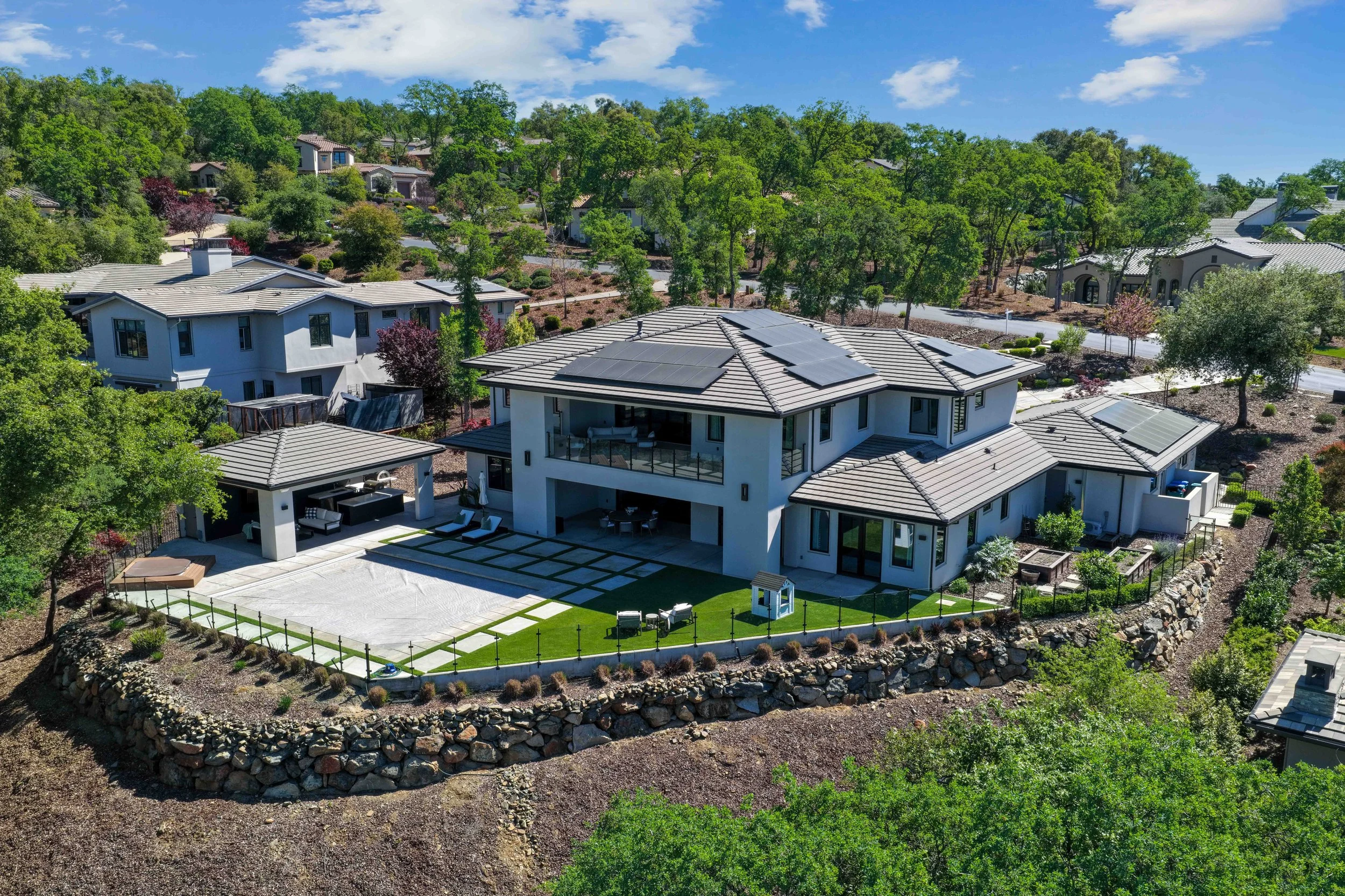 Aerial view of modern house with solar panels, surrounded by trees and landscaped yard, in a suburban neighborhood.