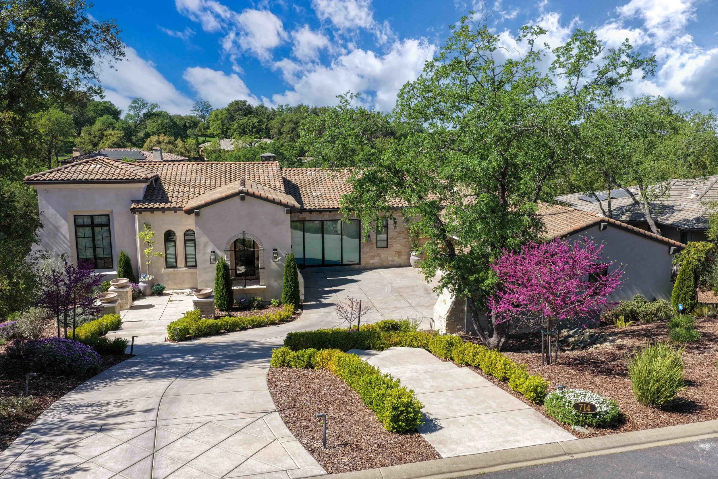 Front yard of a modern house with a tiled roof, a large tree, vibrant pink flowering tree, landscaped garden, concrete walkway, and a house number plaque displaying 714.