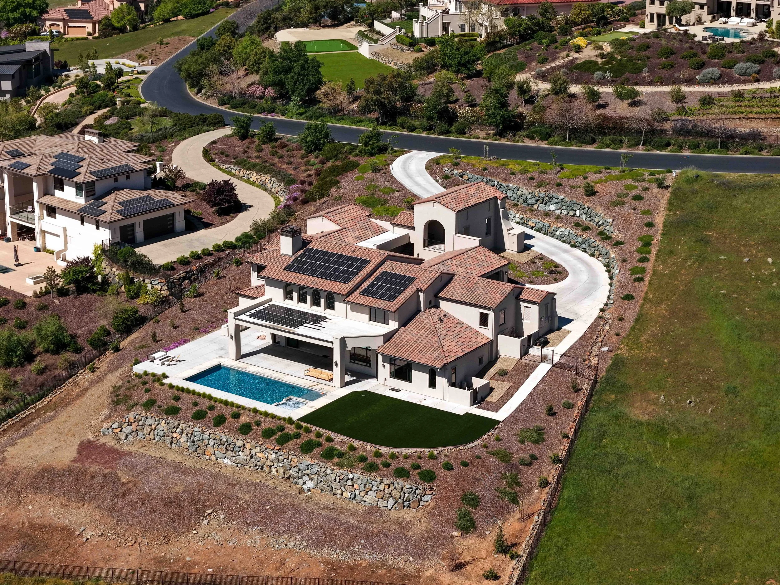 Aerial view of a residential neighborhood with modern houses, some with solar panels on the roofs, surrounded by landscaped yards, roads, and greenery, including trees and shrubs.