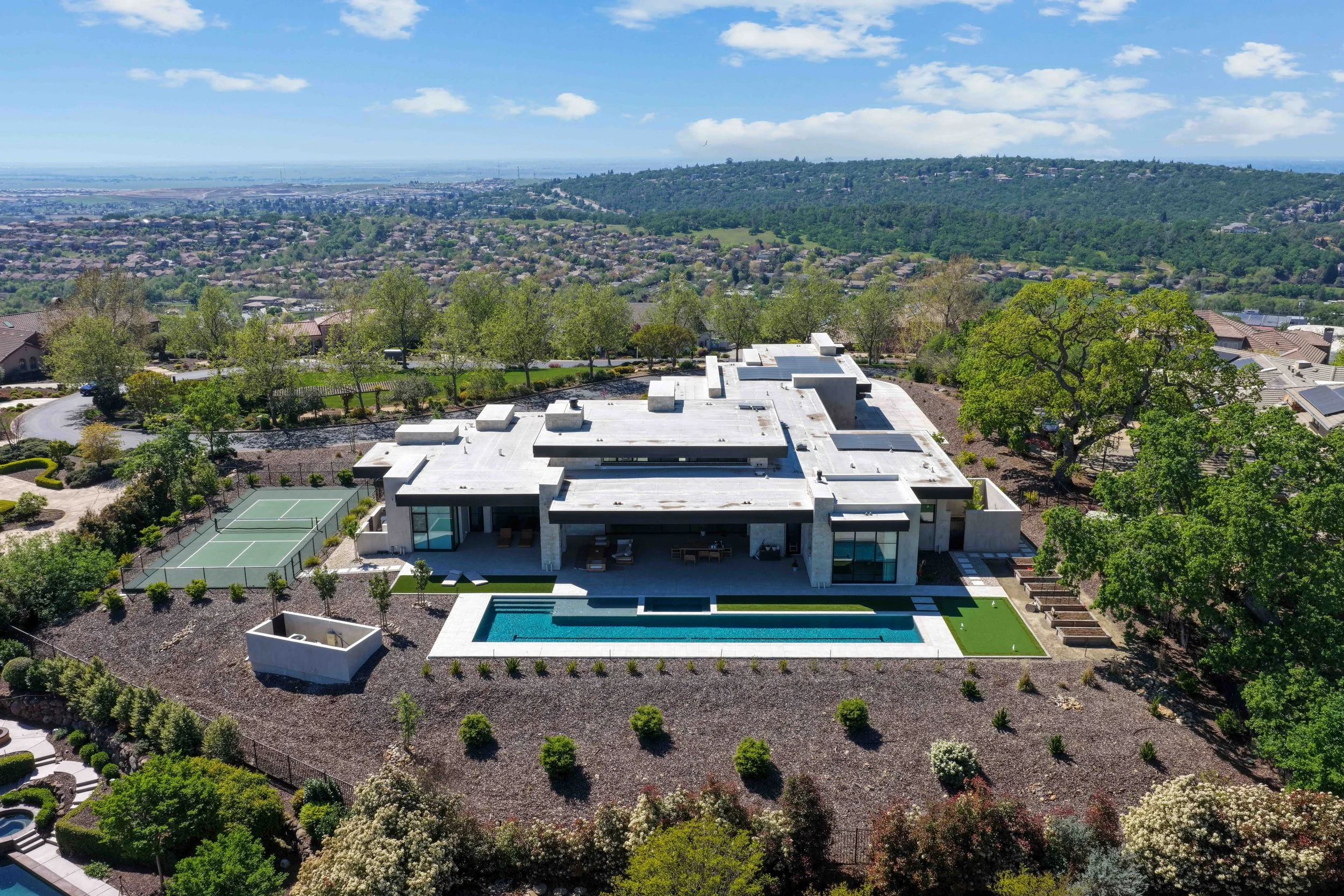 An aerial view of a modern house with a flat white roof, a rectangular swimming pool, and a backyard with a tennis court, surrounded by lush green trees and landscape, overlooking a city and hills in the distance under a partly cloudy sky.