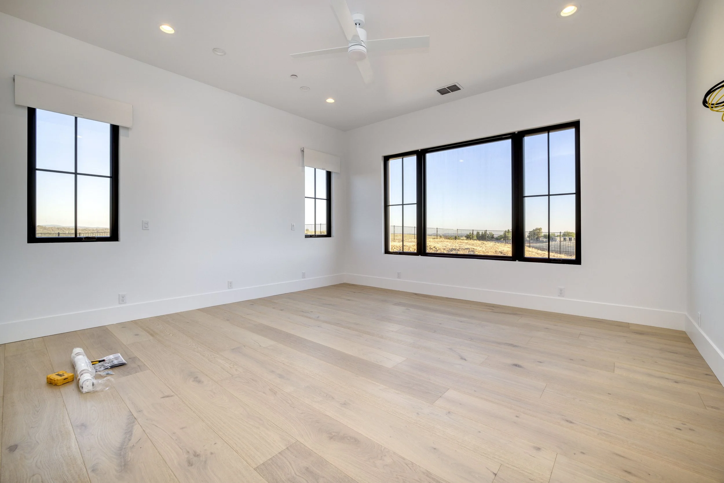Empty room with light-colored wood flooring, white walls, and large windows showing an outdoor landscape. Some construction tools and materials are on the floor.