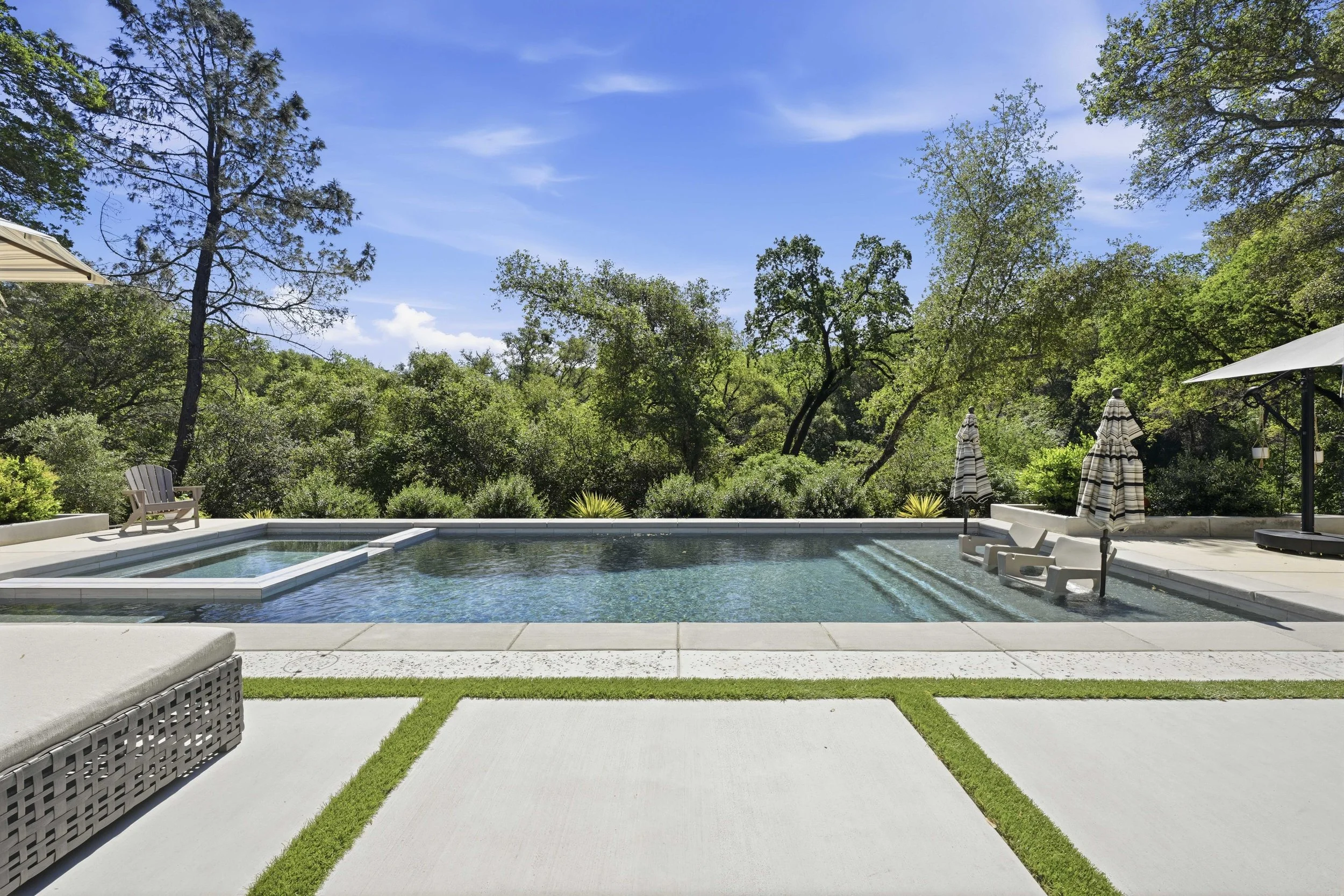 A backyard swimming pool with lounge chairs, umbrellas, and lush green trees under a clear blue sky.