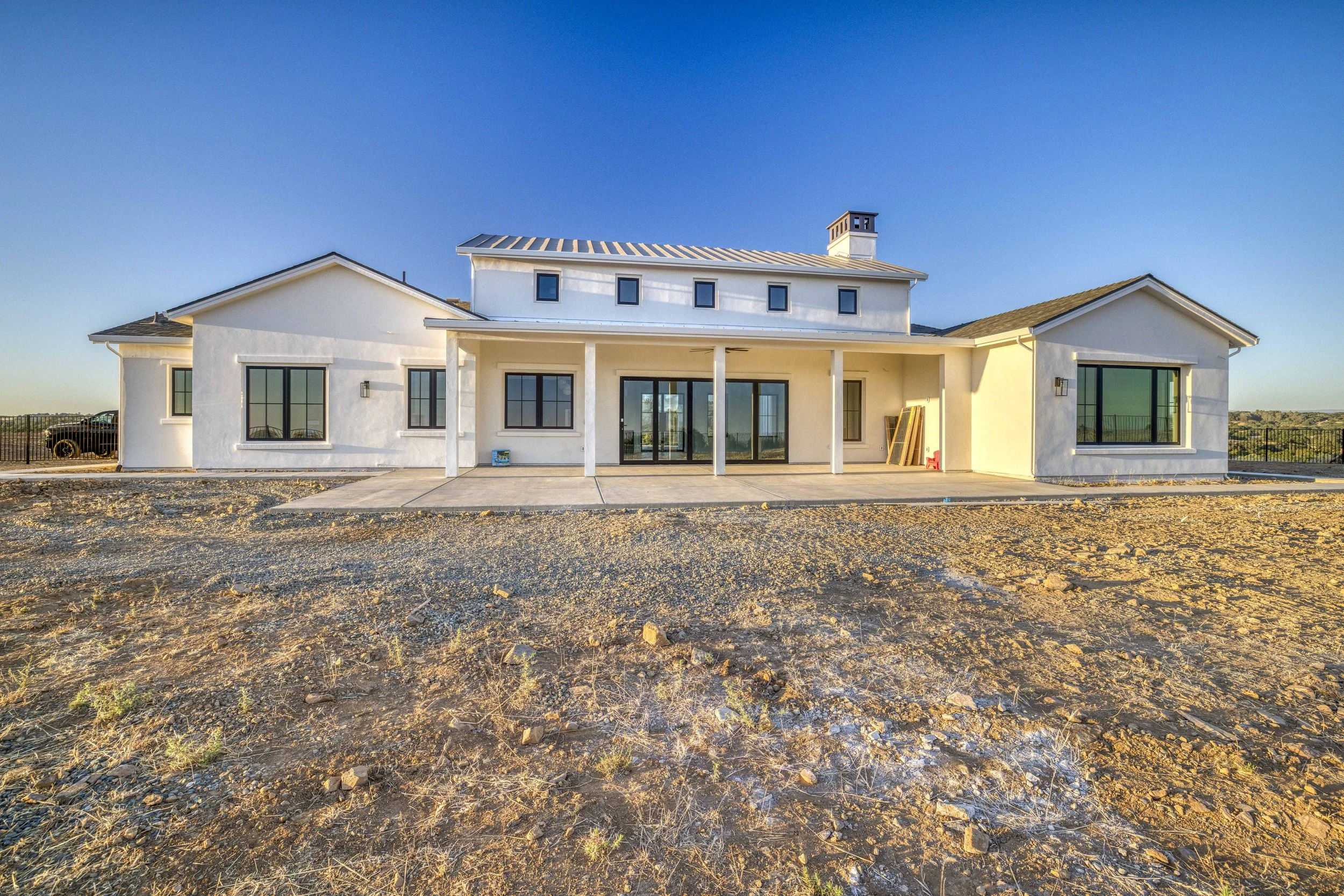 Newly constructed white house with large glass sliding doors and windows, situated on a partly paved and dirt lot under a clear blue sky.
