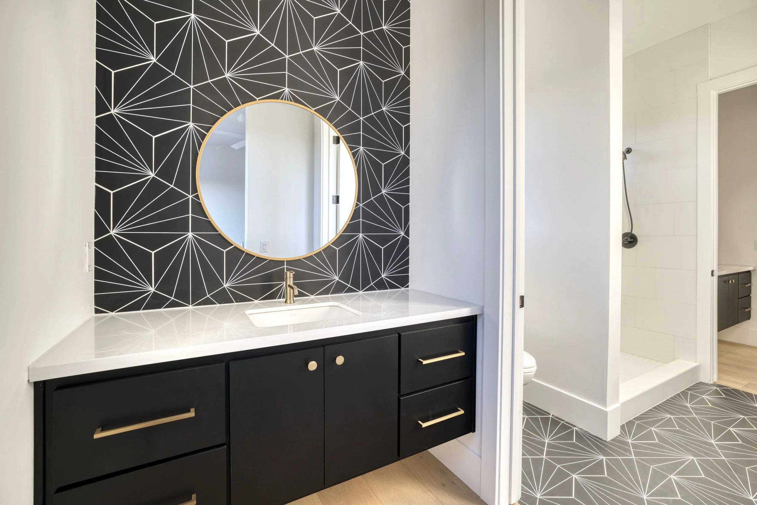 Bathroom with a black and white geometric patterned accent wall, a round mirror with a gold frame, a white countertop with a sink and a gold faucet, black cabinets with gold handles, and a shower area with a handheld showerhead visible through an ope