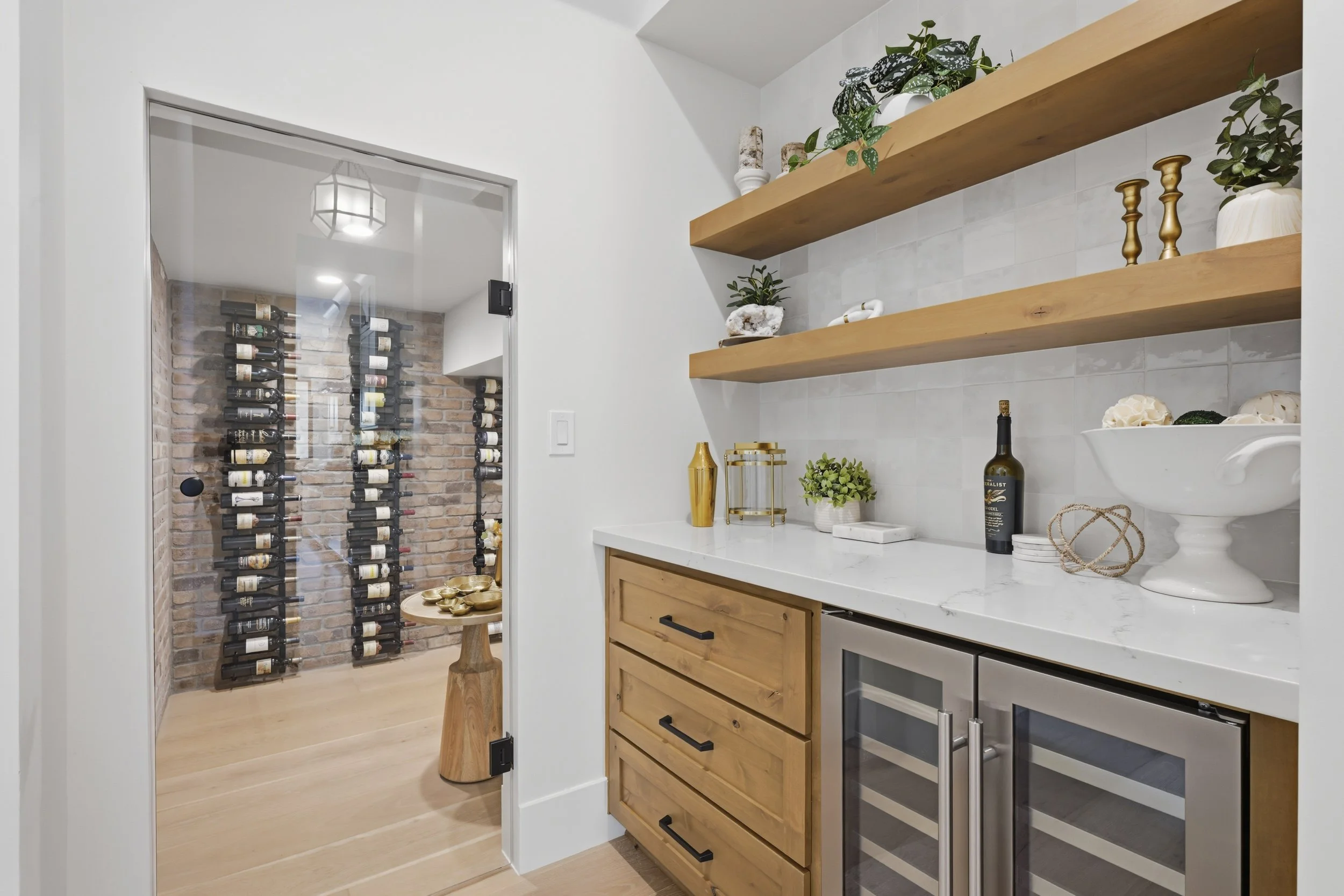 A wine cellar with a glass door and brick wall, featuring multiple rows of wine bottles on racks. Adjacent is a small table with gold bowls. Inside the room, there are stacked wine bottles, a wine cooler, and decorative items on the shelves.