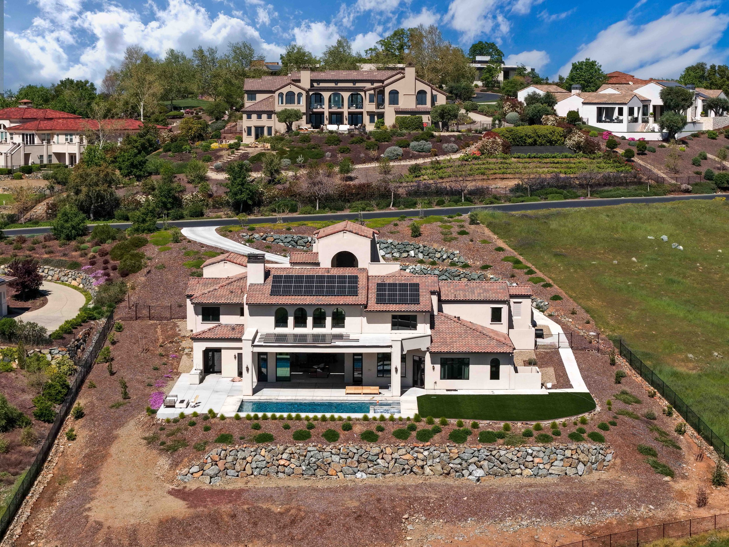 A hillside view showing several houses, with the central one being large and modern, surrounded by landscaped gardens, trees, and a terraced yard. In the foreground, there is a partially built area with a small structure, stones, and a dirt path.