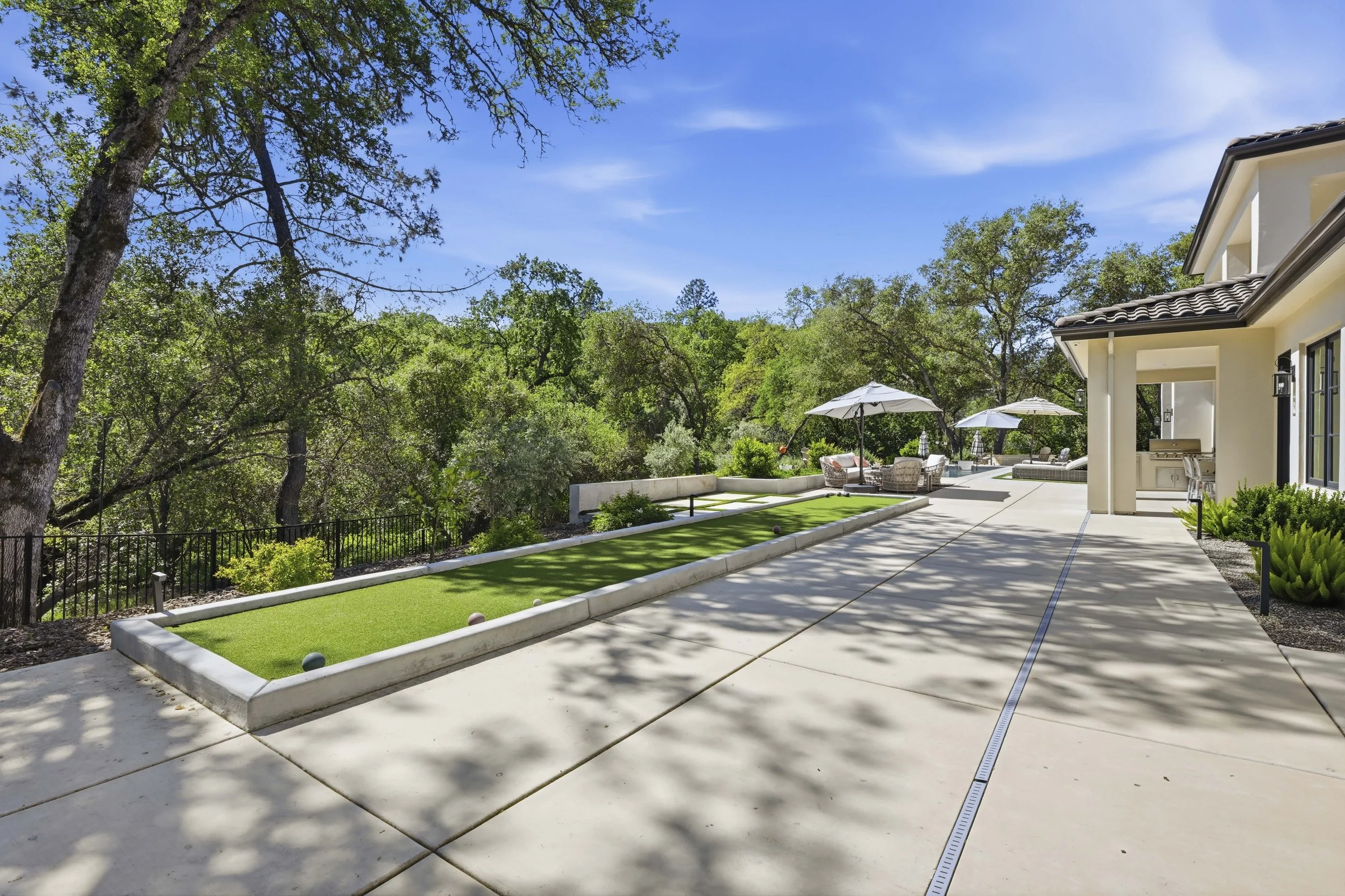 A backyard patio with a putting green game area, surrounded by a garden, patio furniture with umbrellas, and a house with a tiled roof, under a bright blue sky.