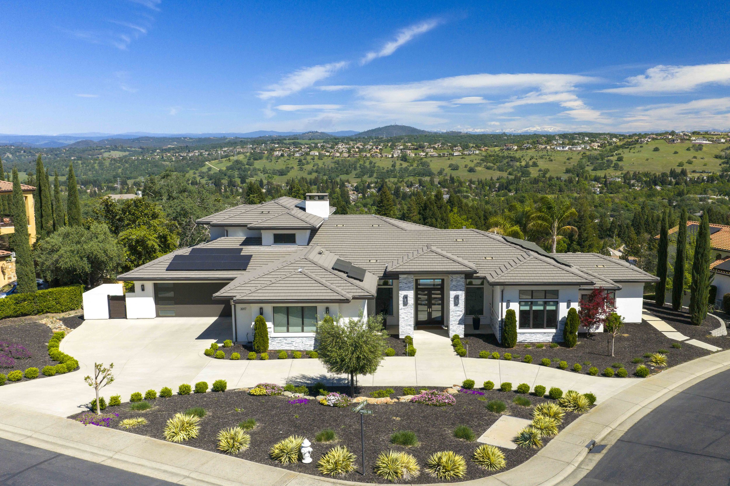 Modern house with a landscaped front yard featuring small bushes, flowers, and palm trees, situated in a suburban neighborhood with rolling hills, green trees, and a bright blue sky in the background.