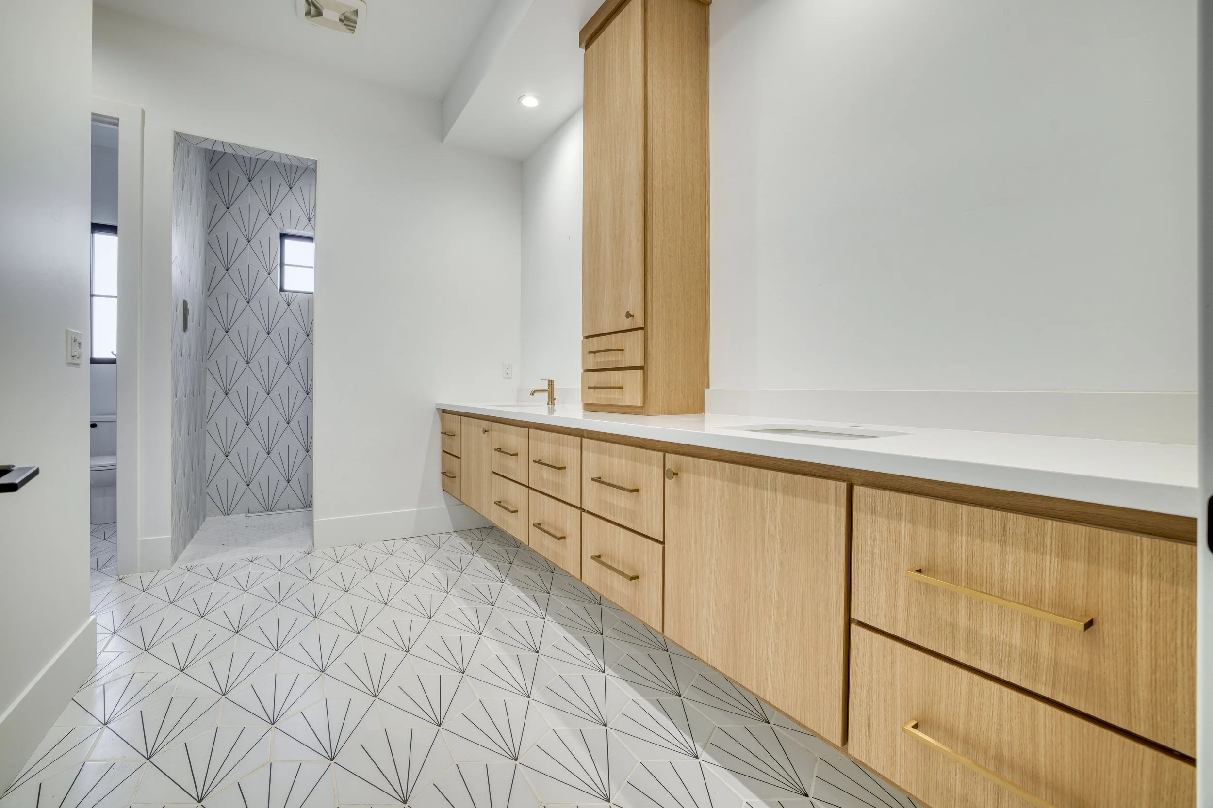 Modern bathroom with a white countertop, wooden cabinets, patterned floor, and a shower with geometric tile design.