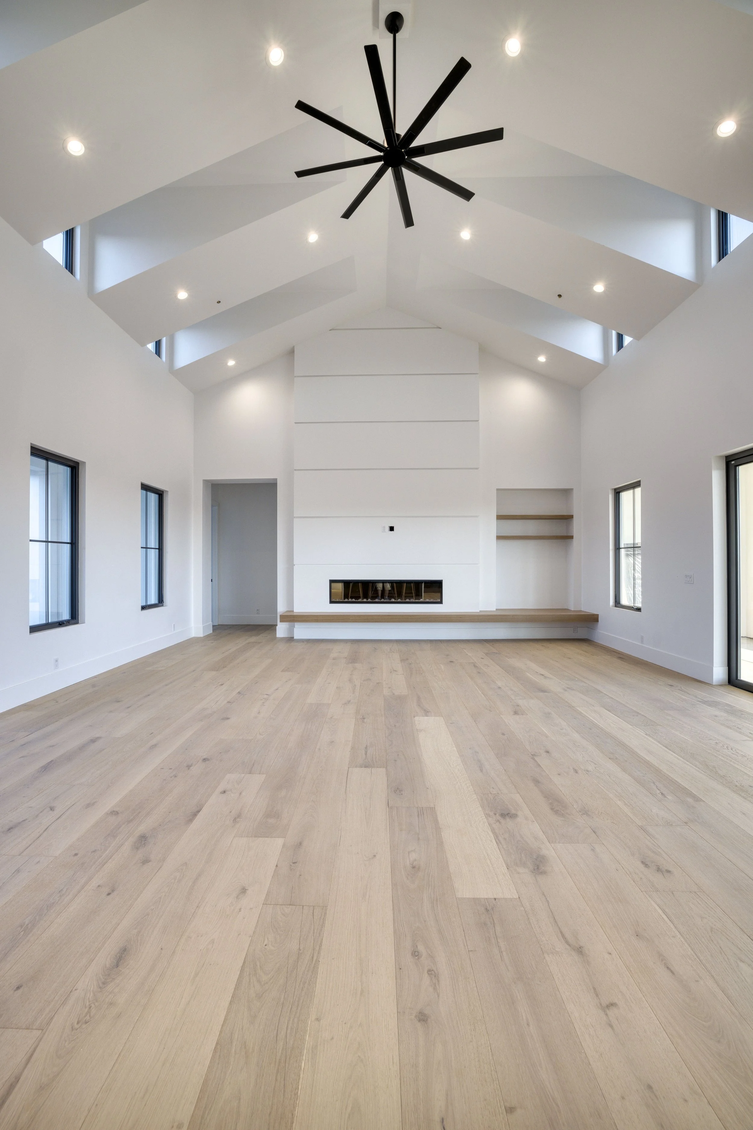 Interior view of a modern, minimalistic living room with high ceiling, skylights, large windows, white walls, hardwood floors, a fireplace, and built-in shelves.