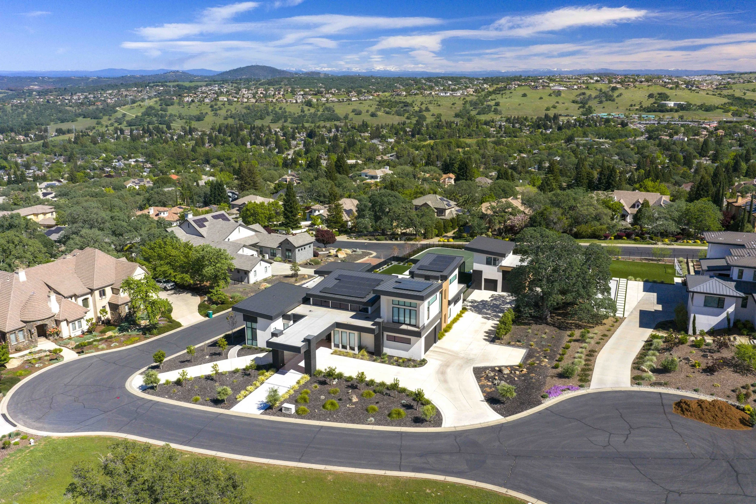 Aerial view of a suburban neighborhood with modern houses, winding streets, and lush greenery under a blue sky with scattered clouds.
