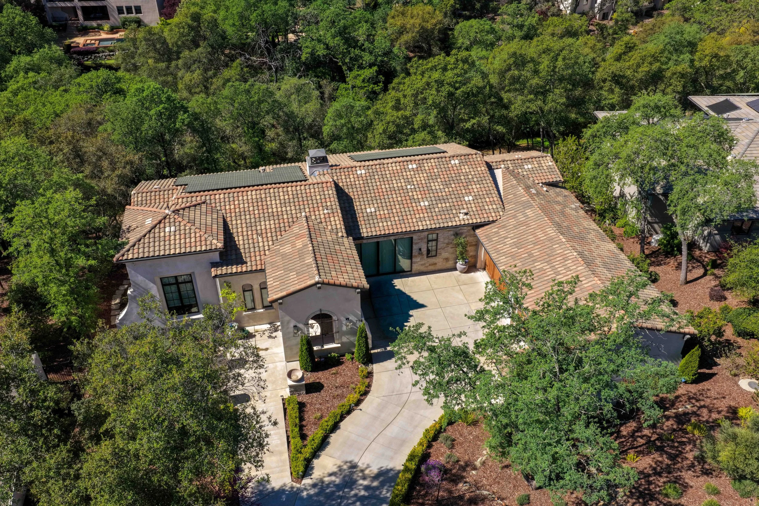 Aerial view of a house with a tiled roof, solar panels, a chimney, and a backyard with trees and a pool, surrounded by lush greenery.
