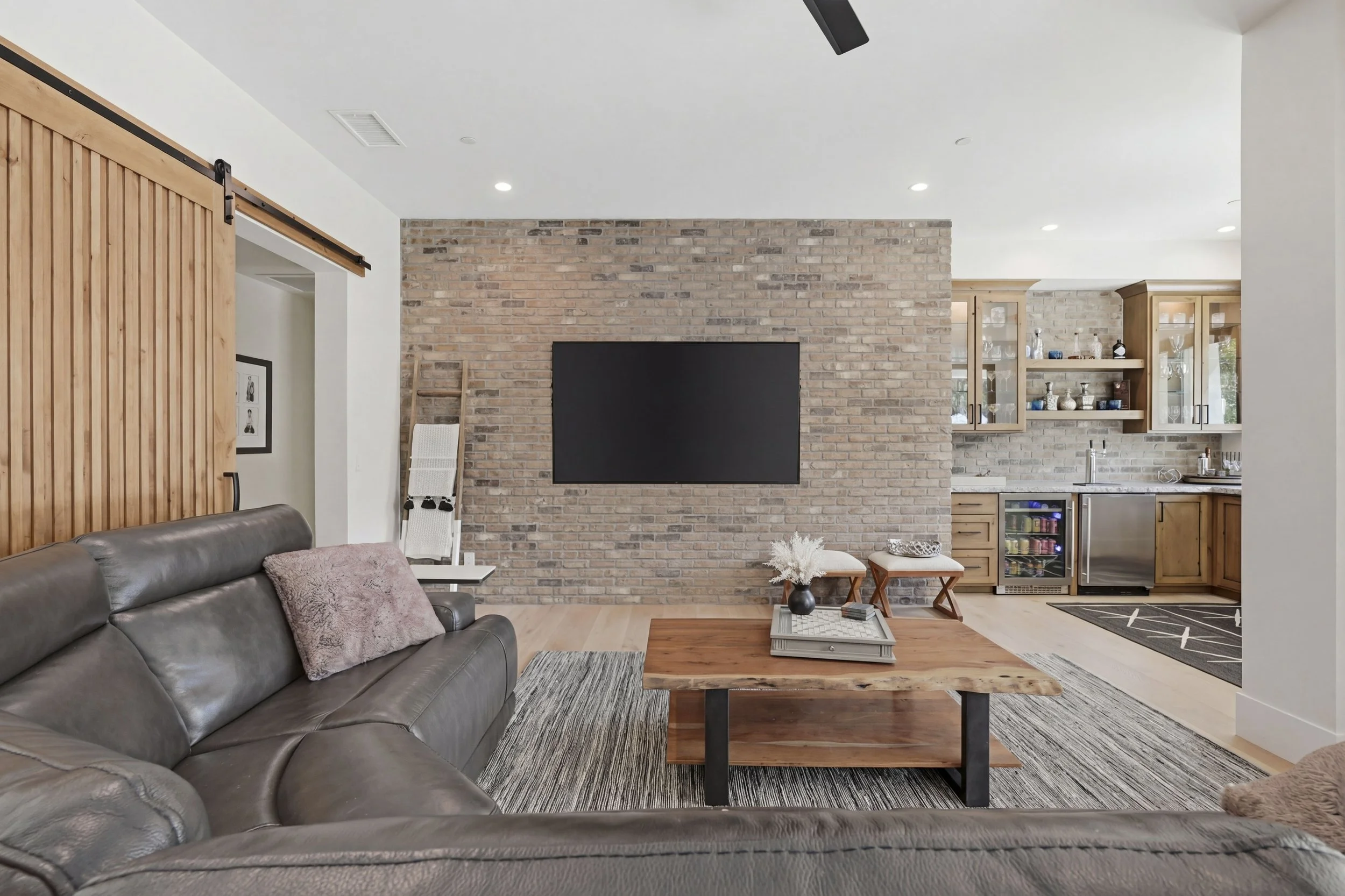 Living room with a brown leather couch, pink pillow, brick wall with a mounted TV, wooden sliding barn door, small coffee table with decorative items, and an open kitchen with wooden cabinets and glass display shelves.