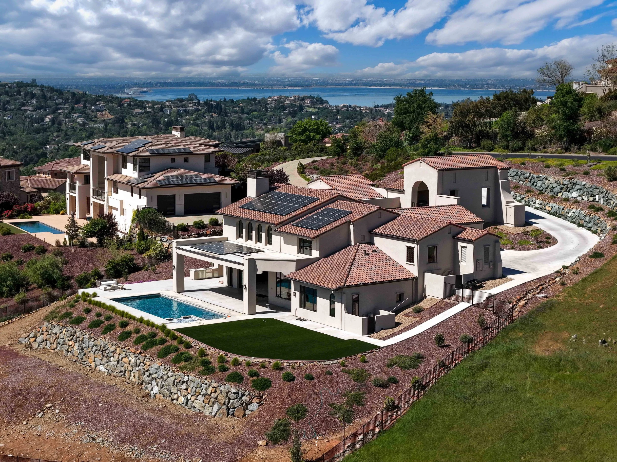 Aerial view of a luxurious white house with solar panels on the red-tiled roof, a backyard swimming pool, and a landscaped yard surrounded by a stone wall in a hilly area with greenery, overlooking distant water and city skyline.