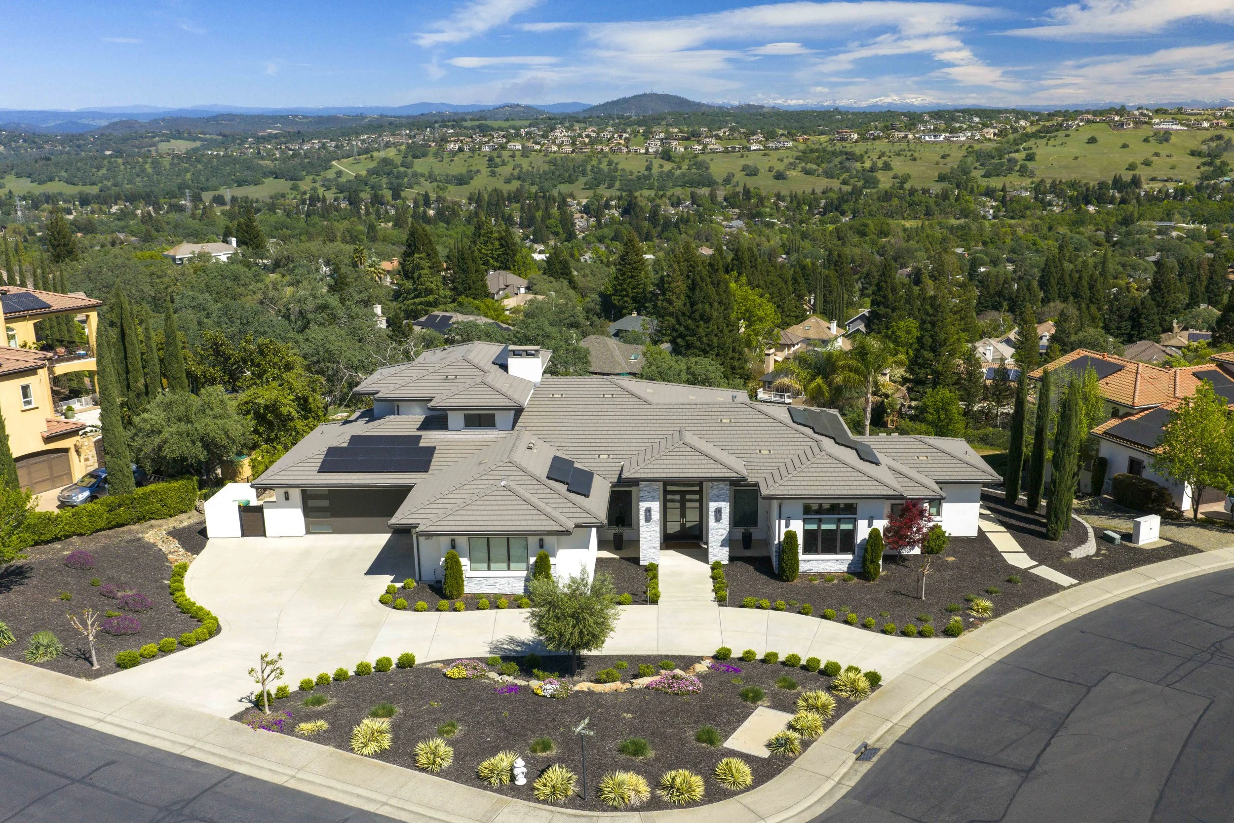 A modern house with a tiled roof, landscaped front yard with plants and trees, and solar panels on the roof, set against a backdrop of a hilly suburban neighborhood under a partly cloudy sky.