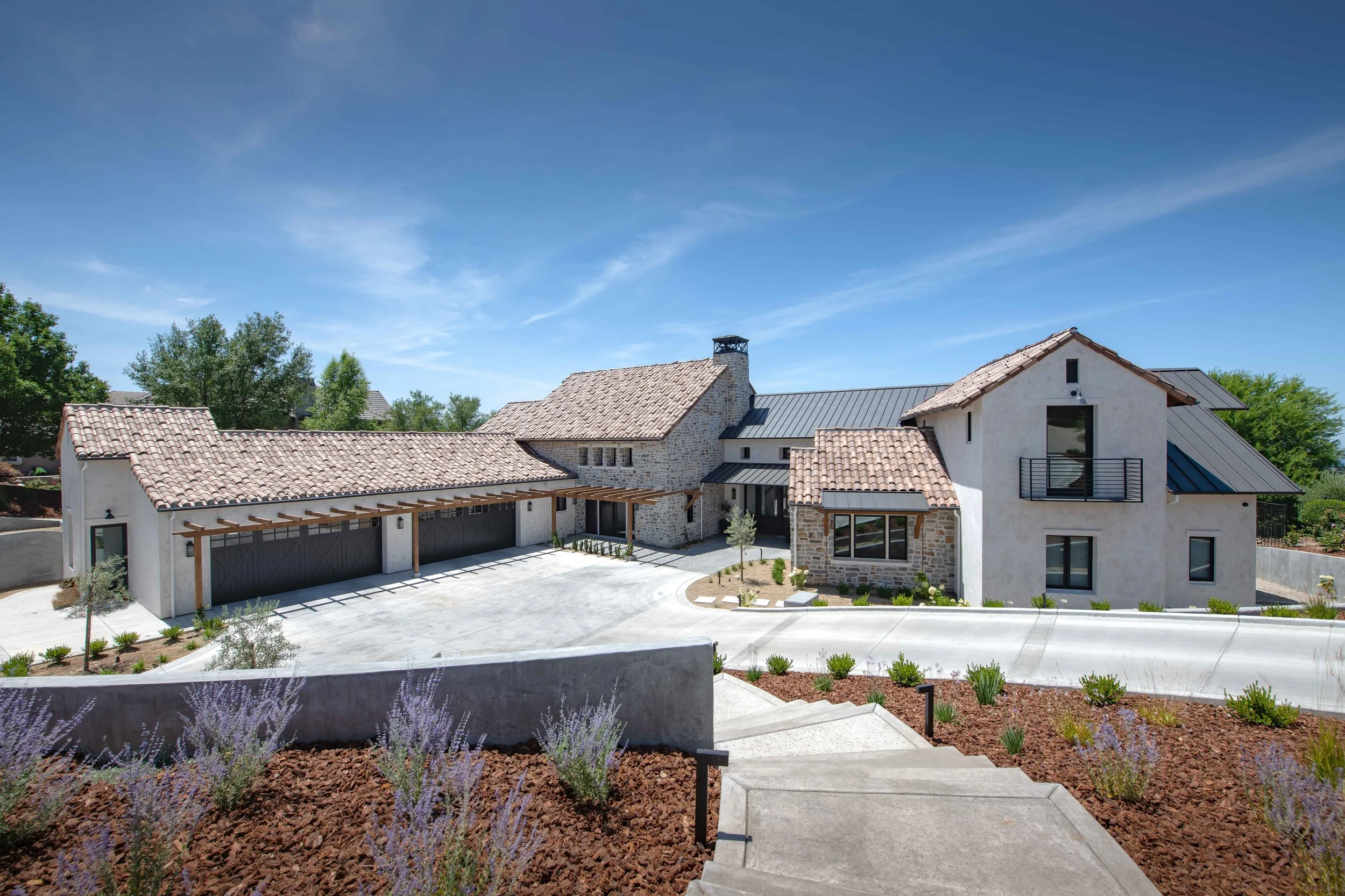 Modern house with tiled roof, stone walls, and a balcony, surrounded by a landscaped yard with plants and a concrete driveway, under a clear blue sky.