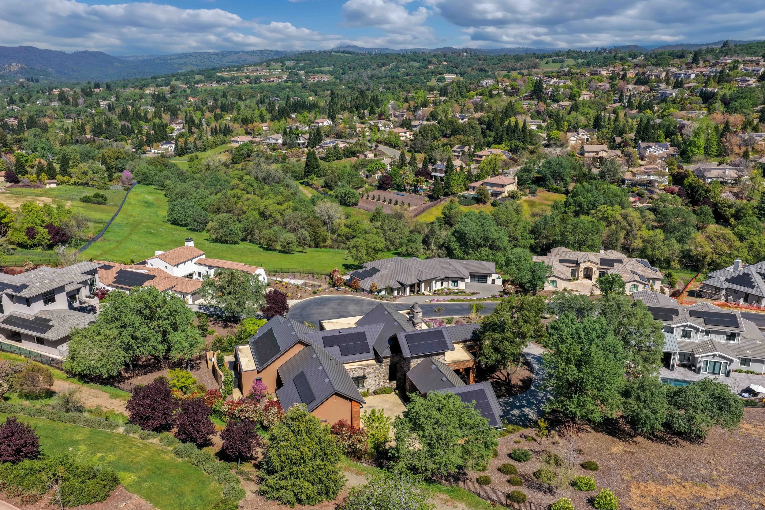 Aerial view of a suburban neighborhood with houses, trees, and green spaces. Rolling hills and mountains in the background under a partly cloudy sky.