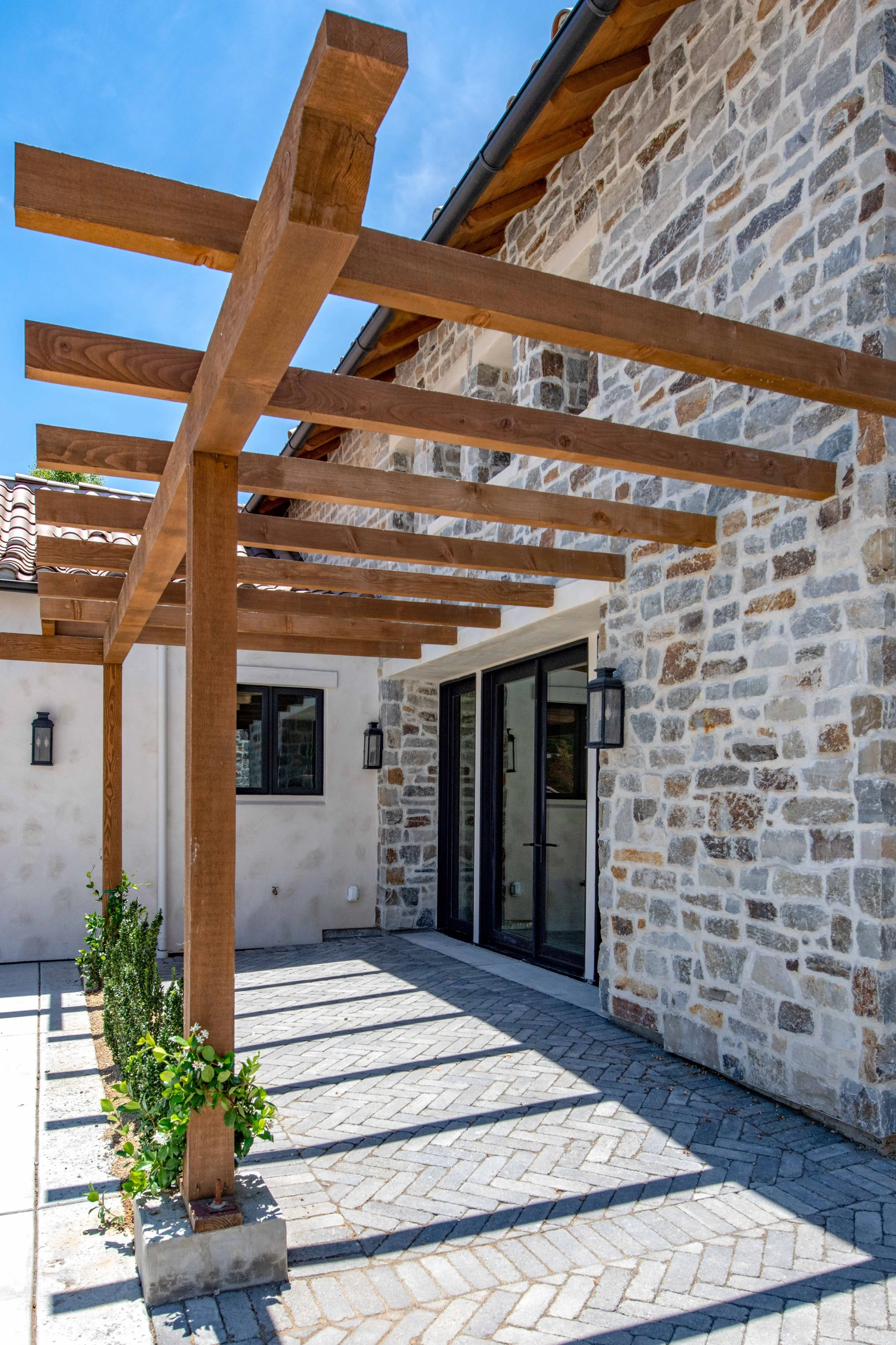 Backyard patio area with a pergola frame made of wooden beams, a stone exterior wall, and a brick paver patio with shadows cast from the pergola. There are some small plants and black outdoor wall lanterns beside a sliding glass door.