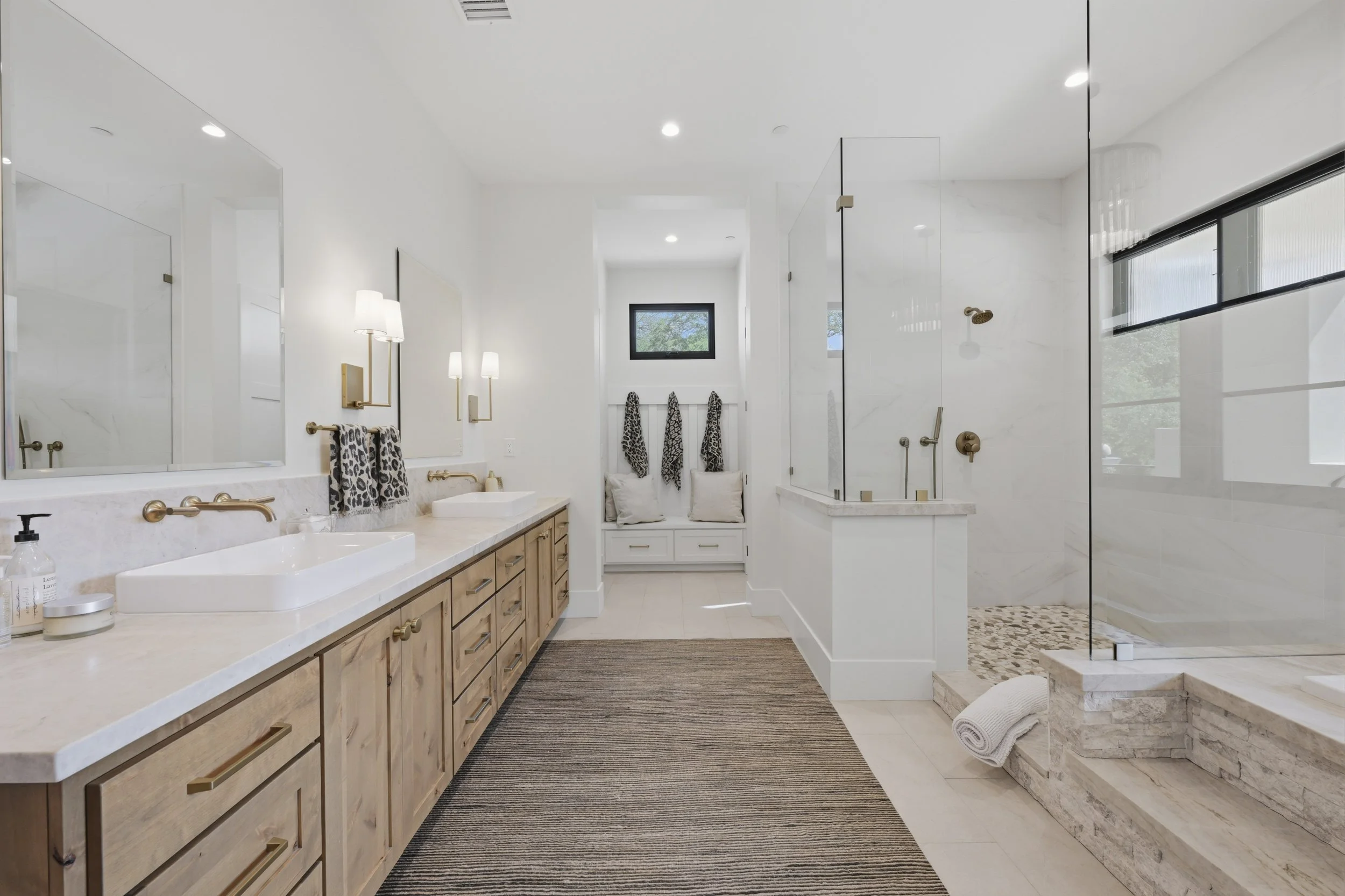 Modern bathroom with white walls and marble accents, featuring a double vanity with wood cabinets, black-framed mirror, and brass fixtures. There is a walk-in shower with glass partition and pebble floor, a window with a seat and leopard print towels