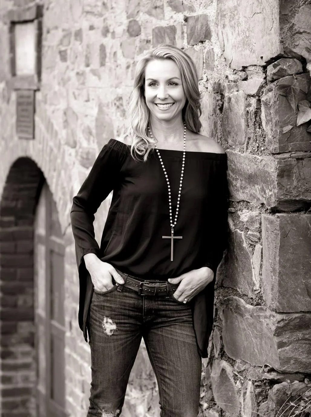 A woman with blonde hair smiling, wearing a black off-the-shoulder top, necklace with a cross, and ripped jeans, standing against a stone wall outdoors.