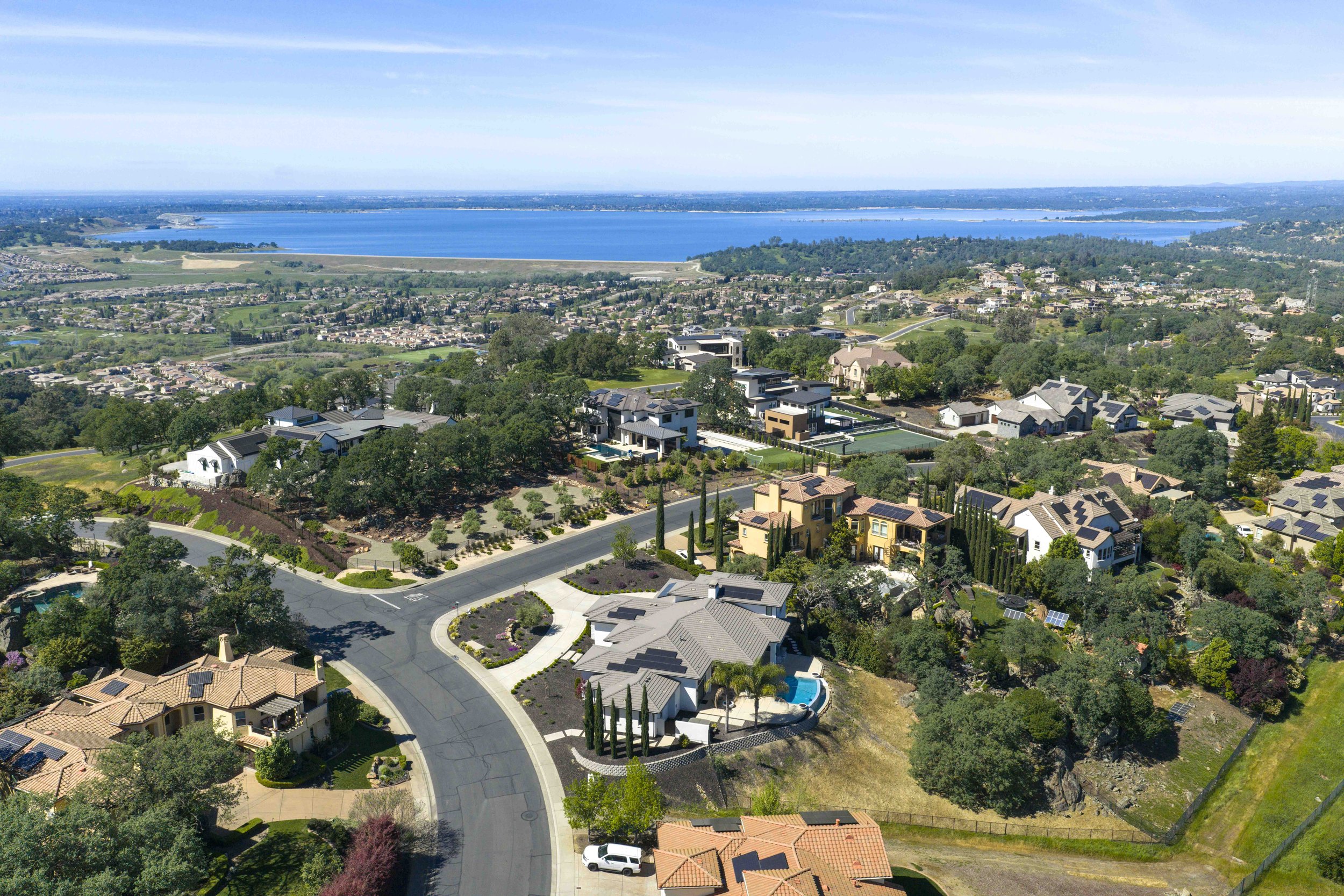 Aerial view of a suburban neighborhood with houses, trees, and streets, overlooking a large blue lake under a clear sky.
