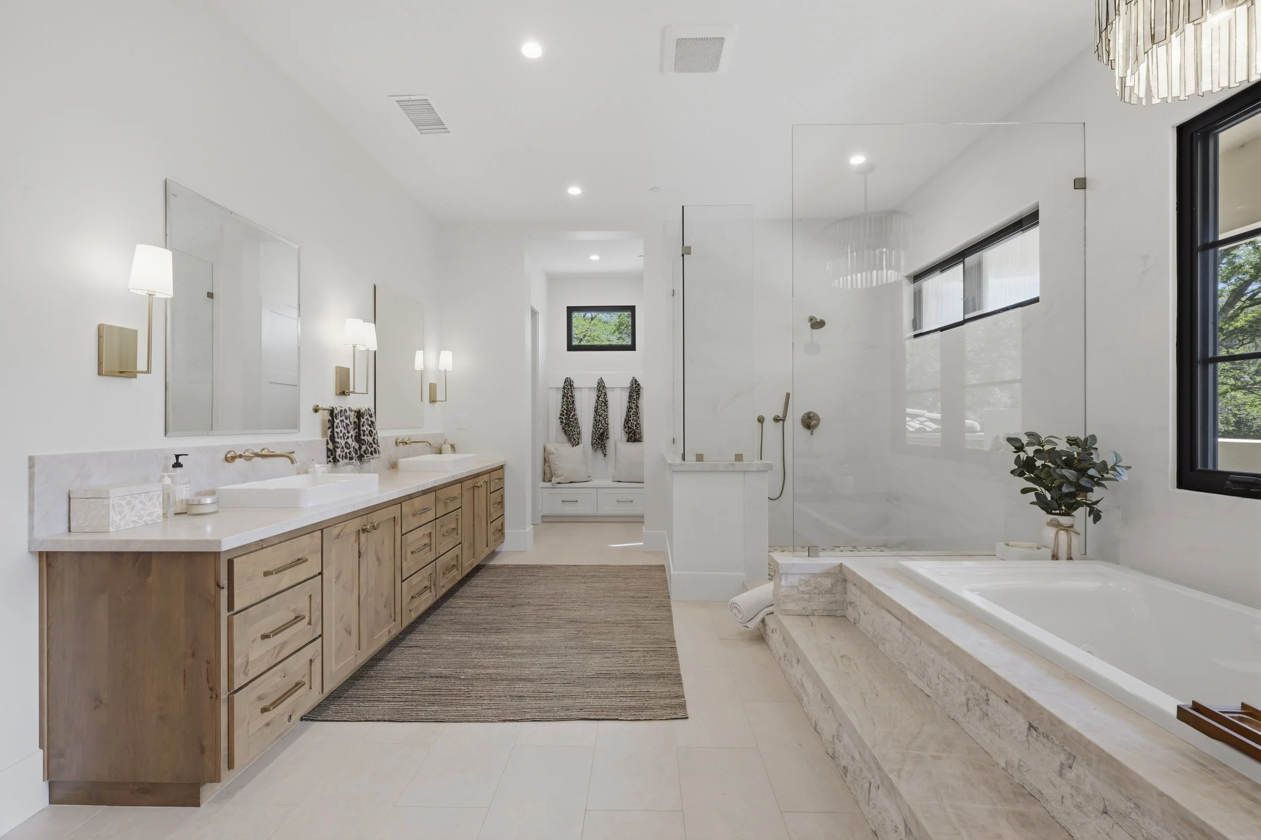 A modern, spacious bathroom with double sinks, wooden cabinet, large mirror, wall-mounted lamps, and a corner shower with glass enclosure. The bathroom features a bathtub with a stone surround, a small potted plant, and large windows for natural ligh