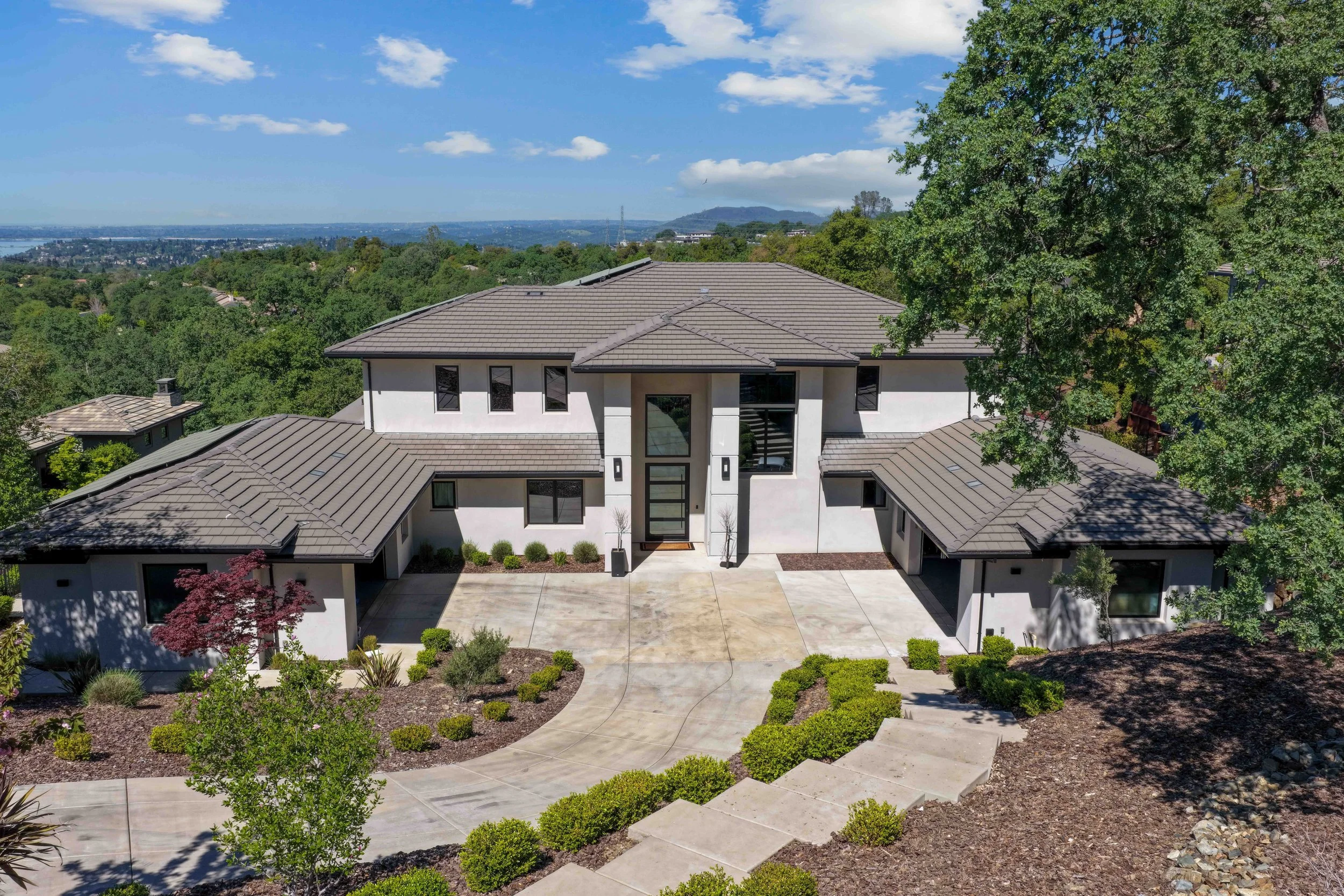 Modern white house with a gray tiled roof, large windows, and surrounding landscaped garden with bushes and small trees, overlooking a distant cityscape and hills under a partly cloudy sky.