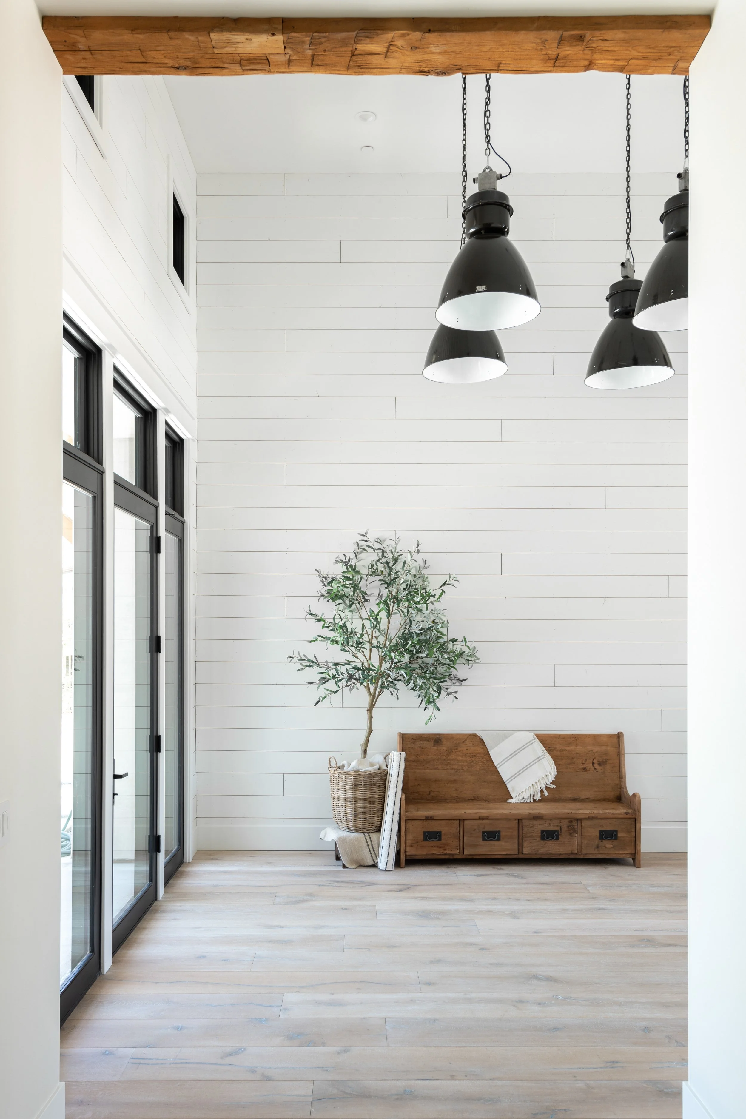 A hallway with wooden ceiling beams, black pendant lights hanging from chains, large windows on the left, a plant in a basket, and a wooden bench with a throw blanket on it against a white shiplap wall.