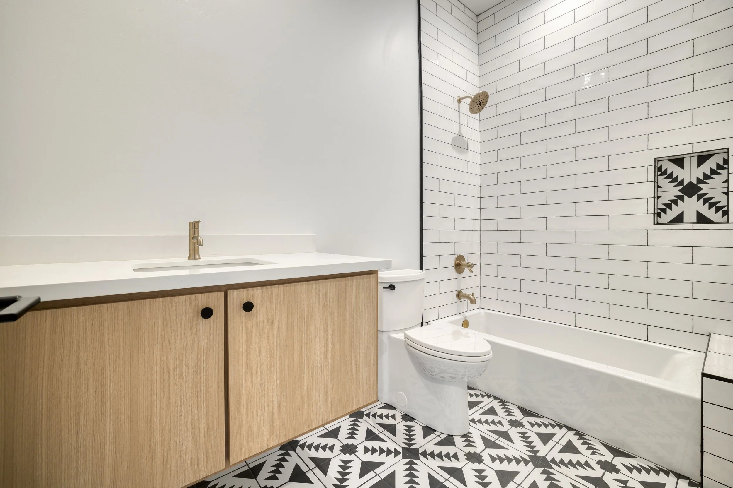 Modern bathroom featuring a white countertop with a built-in sink, wooden vanity cabinet, and brass fixtures. The toilet and bathtub are against a white subway tile wall, with a decorative patterned tile patch near the shower area. The floor has blac