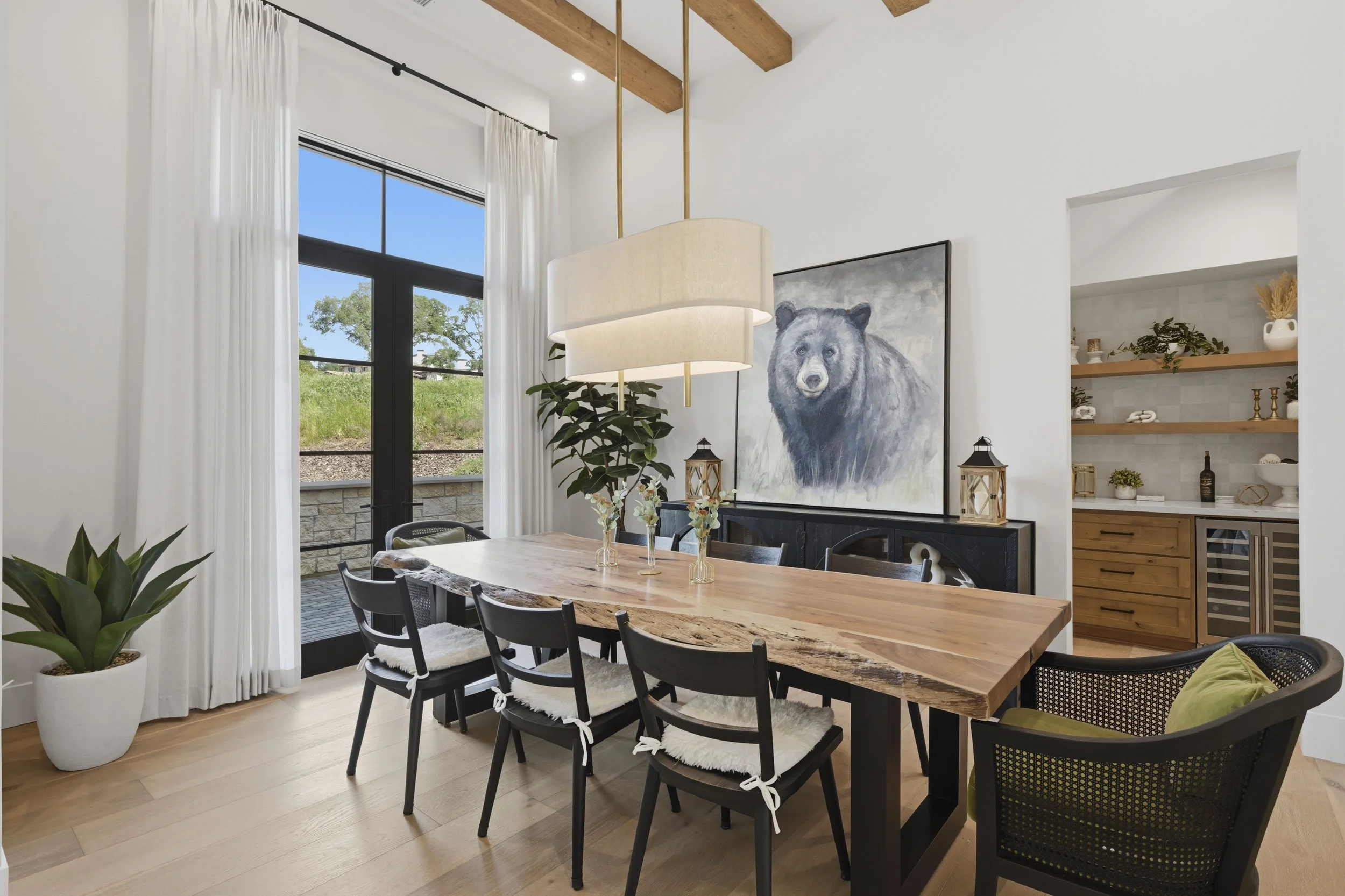 Modern dining room with a wooden table surrounded by black chairs, white curtains, large window showing a green outdoor landscape, and wall art of a bear.