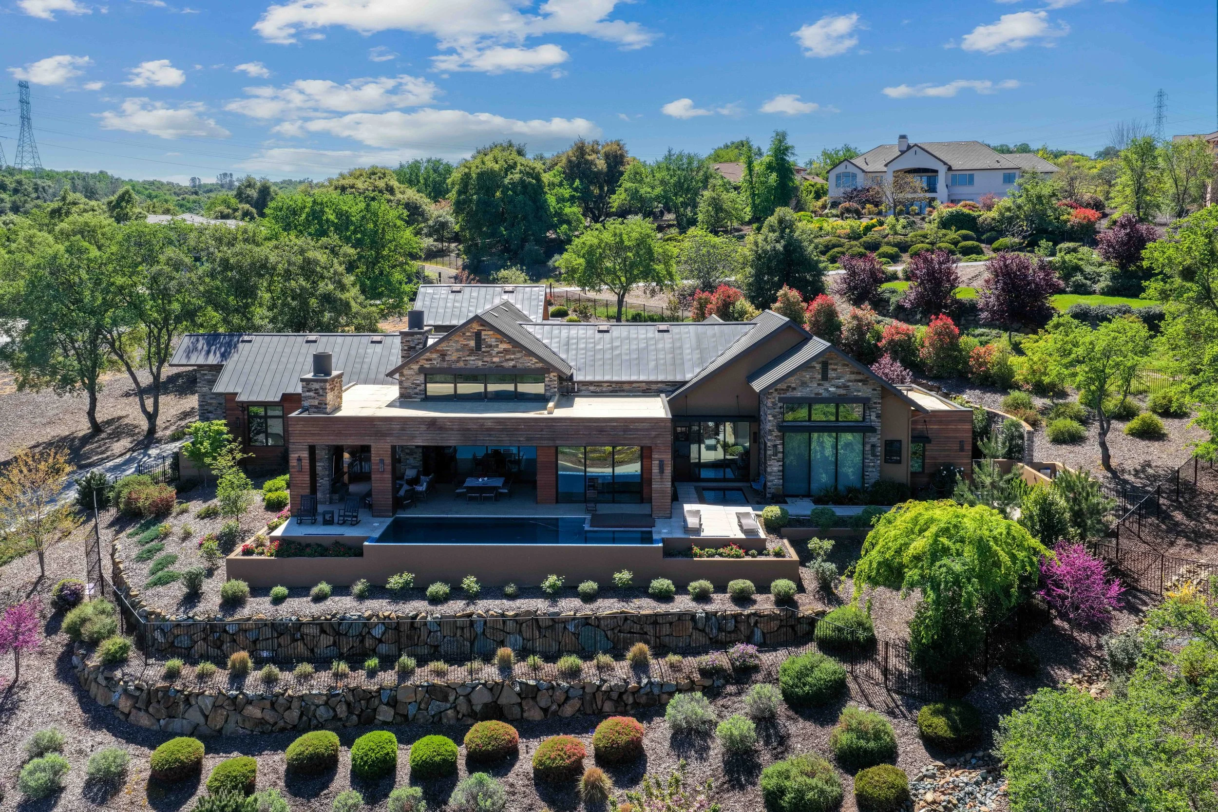 Aerial view of a modern house with a backyard swimming pool surrounded by landscaped garden with trees and flowering bushes, located on a hillside with other houses and green landscape in the background under a partly cloudy sky.