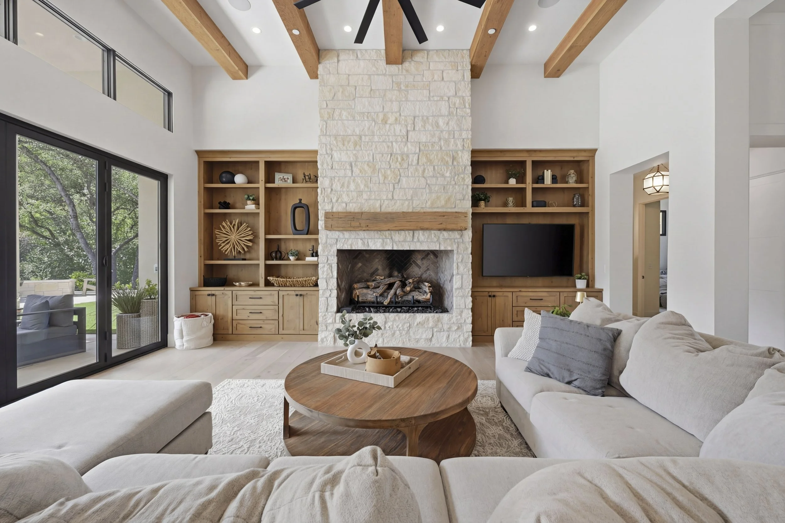 Living room with white sectional sofa, round wooden coffee table, built-in wooden shelves, stone fireplace, large windows, and a ceiling with wooden beams.